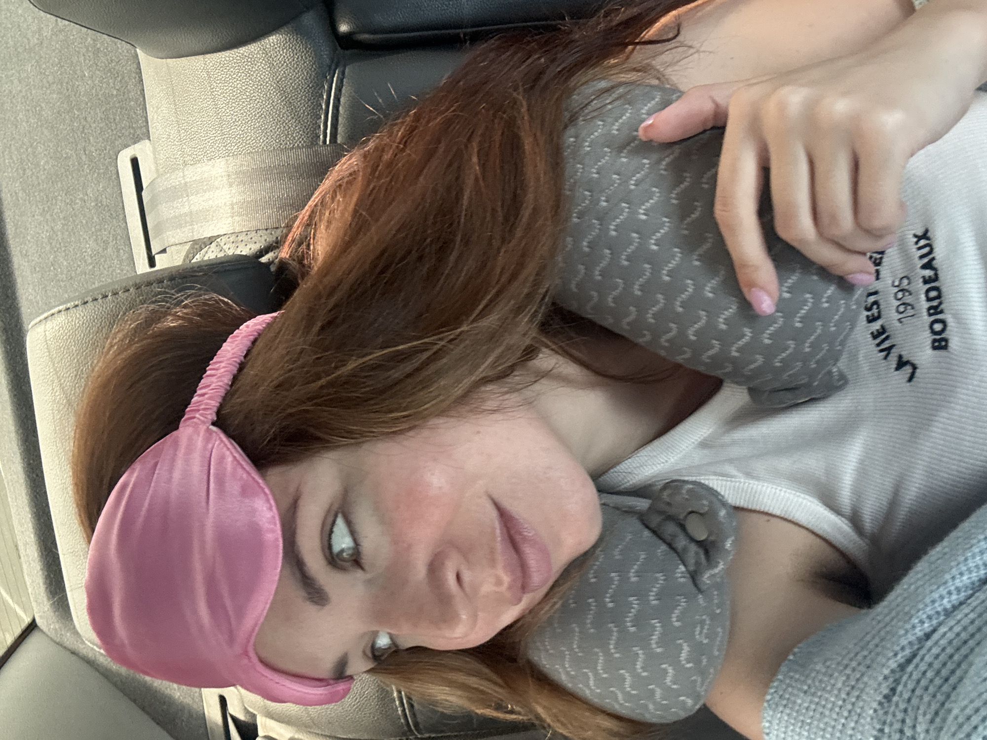 The image shows a woman with long, brown hair wearing a pink sleep mask and a grey patterned neck pillow. She is seated in the back seat of a car, indicated by the black leather upholstery and a seatbelt visible. The woman is looking to her left, with a slight smile. She is wearing a white tank top with text that reads "LA VIE EST 1995 BORDEAUX". Her right hand is resting on the neck pillow. The lighting suggests it is daytime, possibly during travel. The scene does not provide specific landmarks or details to confirm a location within the city of Sychavka, Ukraine.