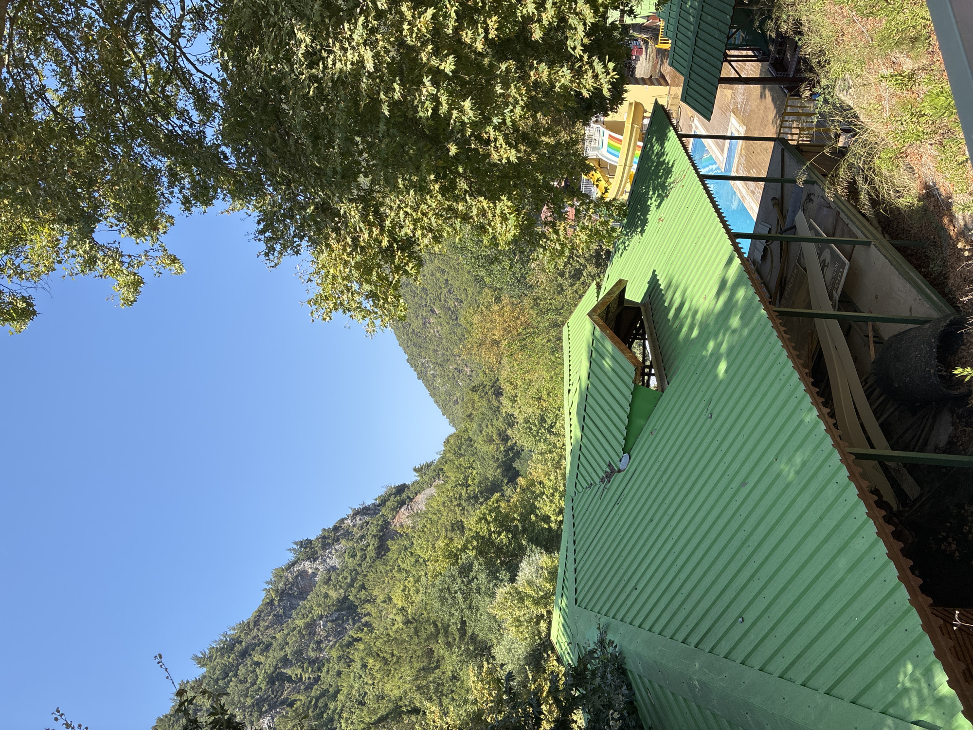 The image shows a view of a resort or park area with a vibrant green, corrugated metal roof in the foreground. Behind the roof, a bright blue swimming pool is visible, and to the right, a yellow water slide with colorful rainbow stripes arcs into the pool. The area is nestled within a lush, green valley surrounded by steep, tree-covered mountains under a clear blue sky. The foliage is dense, with various shades of green indicating different types of trees and vegetation. The overall scene suggests a natural, recreational setting, likely in Alanya, Turkey, given the typical landscape and the presence of such amenities.