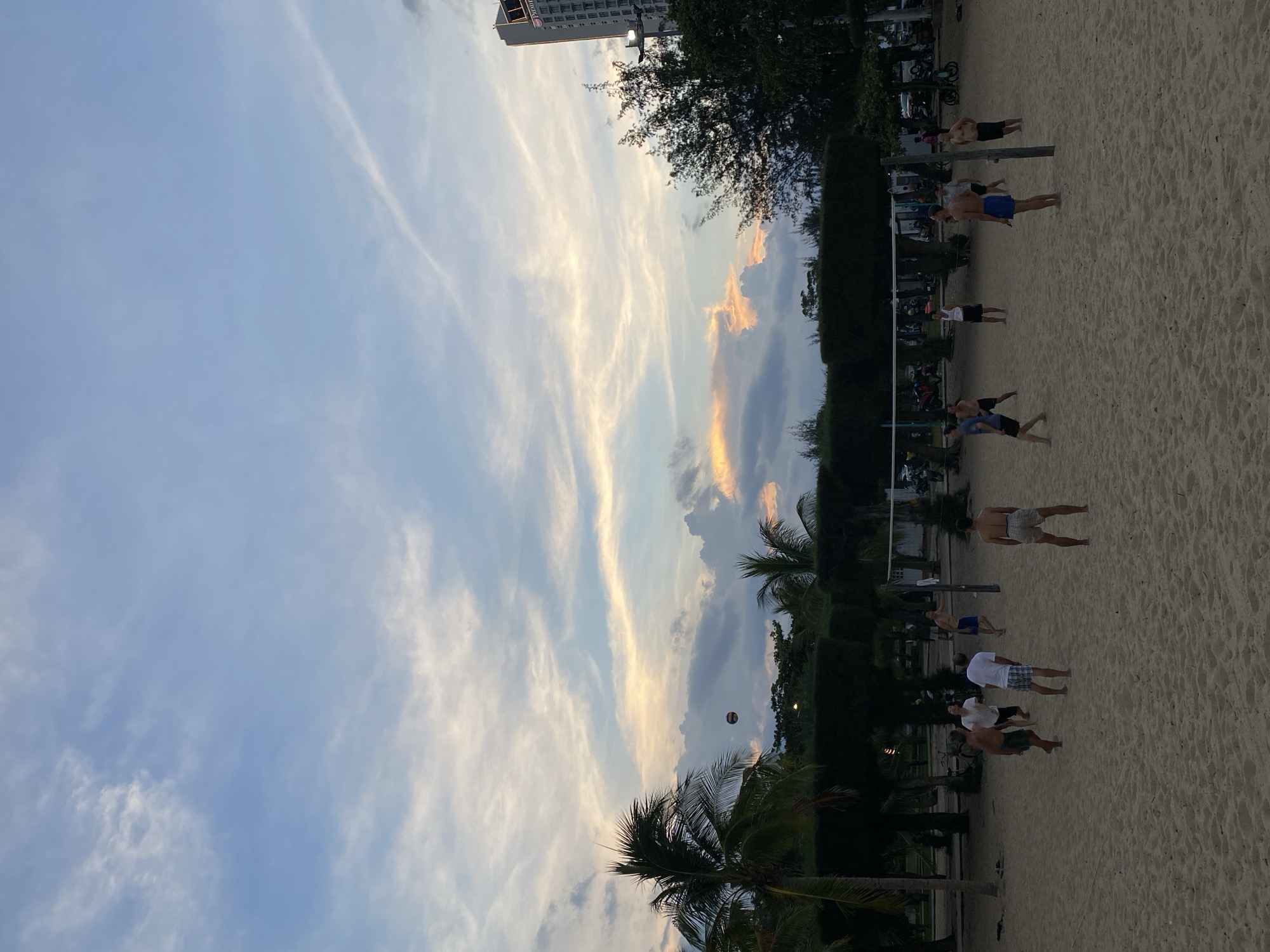 The image depicts a beach volleyball game taking place on a sandy court. Several individuals, predominantly men, are engaged in playing volleyball. The court is equipped with a net. Palm trees and dense foliage line the background, suggesting a tropical or coastal environment. In the distance, a tall building, possibly a hotel, is visible, with a sign that appears to read "Premier." The sky is a mix of blue with scattered clouds, some of which are illuminated with the warm hues of a sunset or sunrise. The scene suggests a recreational activity in an urban coastal setting, consistent with locations like Nha Trang, Vietnam.