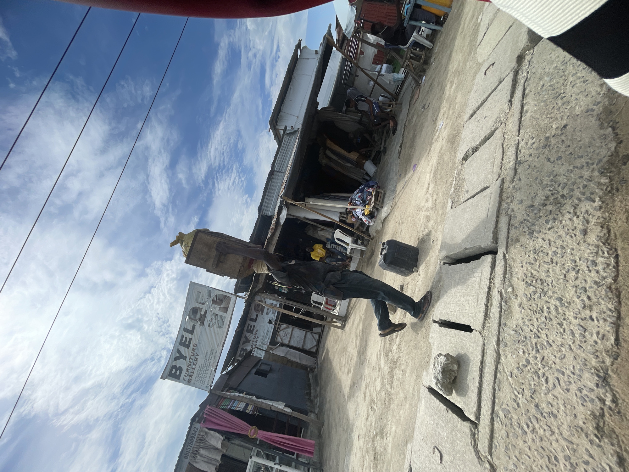 A man in dark clothing is walking along a concrete surface in Ado, Nigeria. He is carrying a wooden object on his head and has a black container on the ground near him. A sign for "BYELO FURNITURE GALLERY" is visible behind him, indicating a local business. The surrounding environment shows a street scene with buildings constructed of various materials, including corrugated metal. Overhead electrical wires and a partly cloudy sky are present. The photo appears to be taken from a low angle.