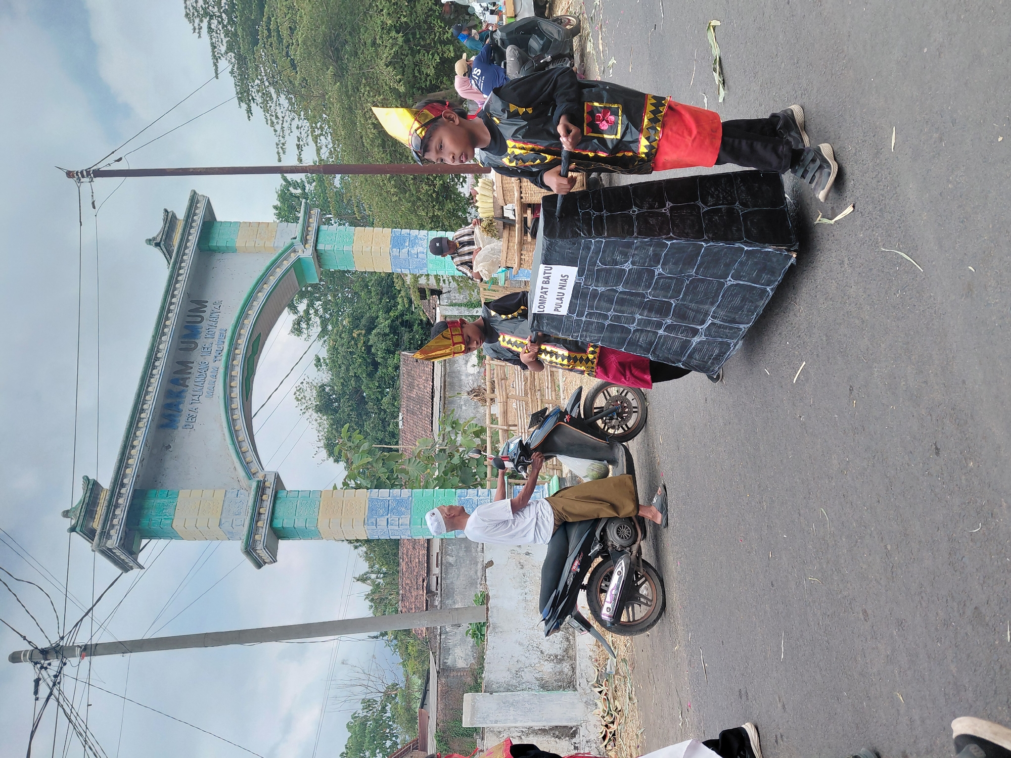 In the city of Bimo, Indonesia, a street scene unfolds beneath an overcast sky near an archway that marks the entrance to a cemetery labeled "MAKAM UMUM DESA TALIANAMPE KEC. KOTA ANYAR MAKAM THUWER". Two young boys, dressed in elaborate black and red costumes with gold accents and conical headdresses, are positioned in the foreground. They are participating in what appears to be a parade or cultural event, as they are pushing a large, dark, rectangular prop designed to resemble a stone structure with a white label that reads "LOMPAT BATU PULAU NIAS". Behind them, an older man wearing a white cap and a white shirt over a brown sarong is seated on a black motorcycle. The environment includes a paved road with scattered debris, a white concrete wall, and lush green trees. Several other individuals are visible in the background, including a person on another motorcycle and others near a cart laden with corn husks. The overall impression is of a community gathering or celebration.