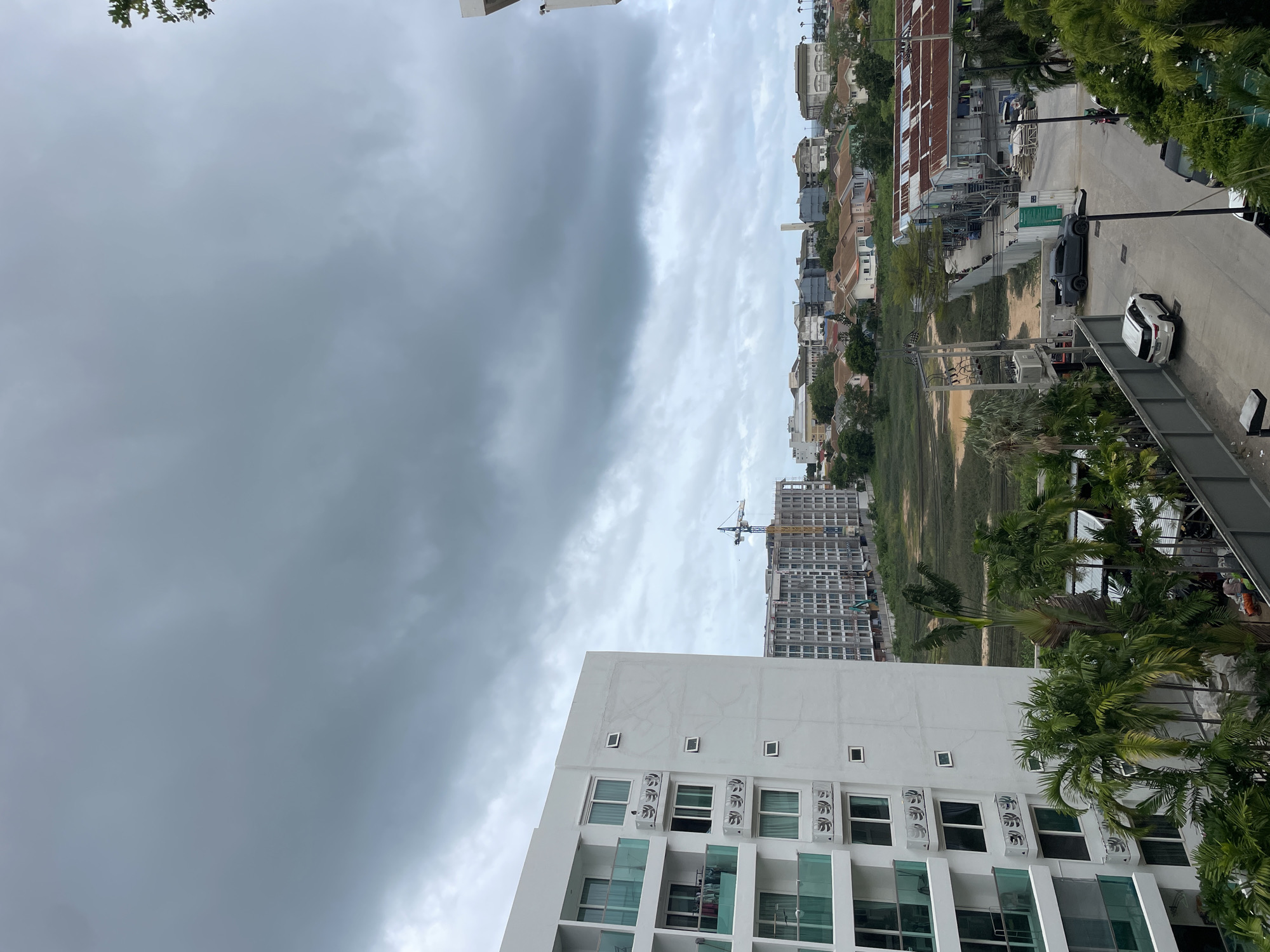 A high-angle, outdoor, daytime, wide shot captures a cloudy, overcast sky above a city landscape in Pattaya City, Thailand. A large, white, multi-story apartment building with balconies and glass railings dominates the left side of the frame. To the right of the apartment building, a mid-rise residential building under construction is visible, with a crane above it. In the middle ground, a collection of houses with red tile roofs forms a suburban-like neighborhood. Further to the right, industrial-style buildings with corrugated metal roofs are present, adjacent to a street. Several vehicles, including a white SUV, a dark grey pickup truck, and a parked motorcycle, are visible on the street. Palm trees and other greenery are scattered throughout the scene, indicating a tropical environment. The overall impression is of a developing urban area under a somber sky.