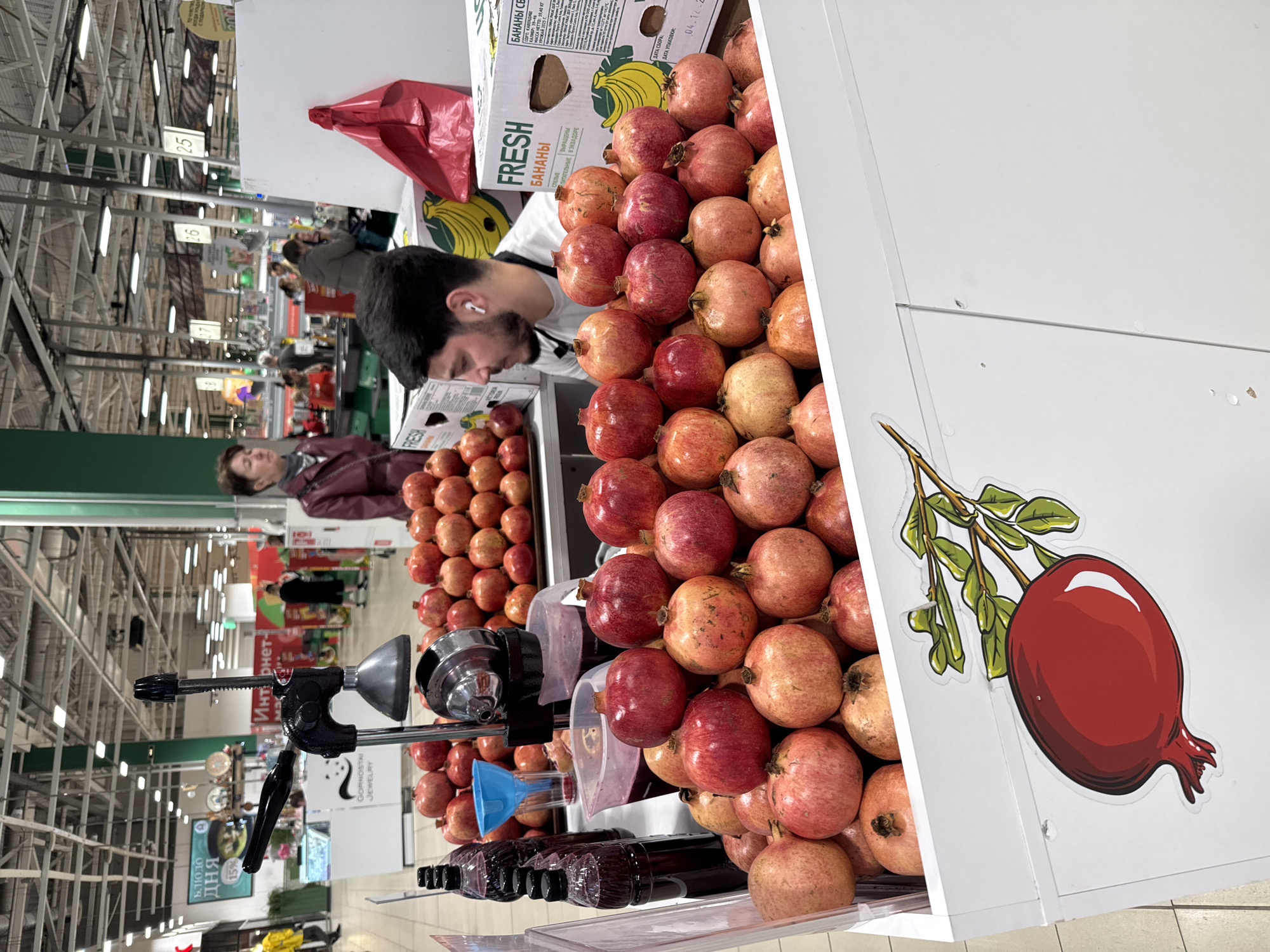 A man wearing an earbud stands behind a display of pomegranates and a manual fruit press, seemingly preparing pomegranate juice. Bottles of juice are visible on the shelf below. A woman wearing a maroon jacket stands in the background. The display is labeled with a sticker of a pomegranate and a box of bananas is visible in the background. The location appears to be a supermarket, likely in Vsevolozhsk, Russia, judging by the Cyrillic text on the packaging and signage.