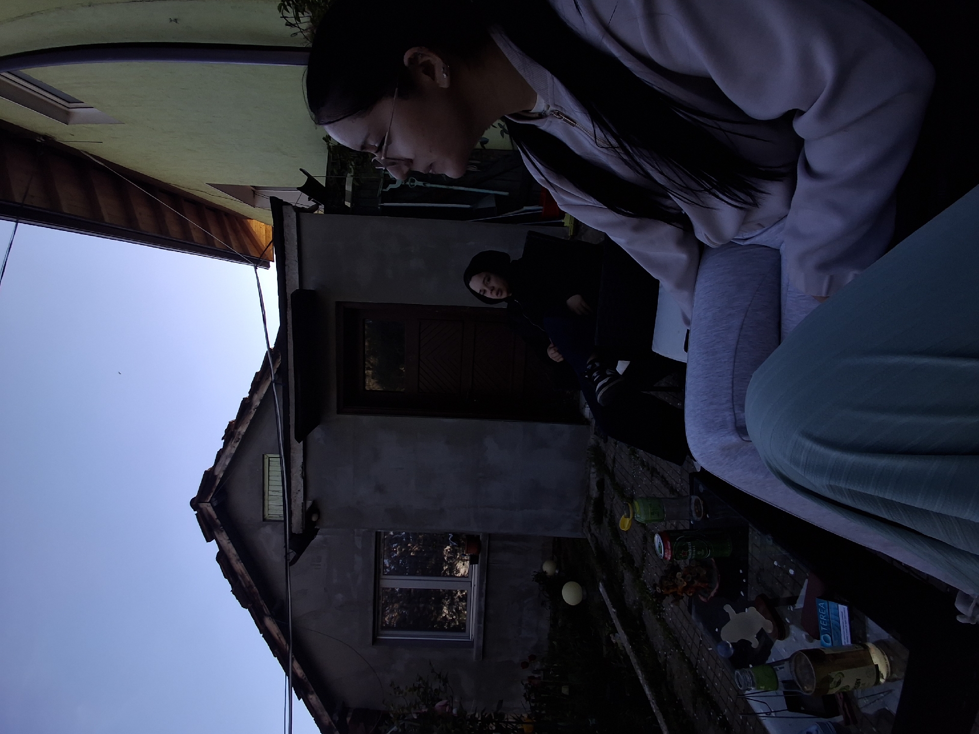 This image, taken in Buhovo, Bulgaria, captures an outdoor evening scene under a clear, dim blue sky. The original image appears to be rotated 90 degrees clockwise.
In the foreground, a woman with long dark hair and glasses is seated, looking downwards. She is wearing a light-colored sweatshirt and grey sweatpants. Part of another person's leg or clothing, in a light blue-green fabric, is visible in the bottom right, suggesting close proximity to the main subject.
Behind the woman, a house with a dark, possibly concrete, facade, a wooden door, and a single window is visible. Another individual, dressed in a dark hoodie, stands partially obscured near the house's door, also looking towards the foreground. To the left, the eaves and light green exterior of an adjacent building are seen against the sky.
An outdoor table occupies the lower right portion of the frame, cluttered with various items. These include several glass jars, metal cans, and organic waste like food scraps. A small blue box with the word "TEREA" clearly printed on it is prominently displayed on the table. Several spherical, possibly decorative, lights are placed on the ground near the table. The soft, fading light indicates dusk, lending a quiet atmosphere to what appears to be a casual backyard or patio setting.