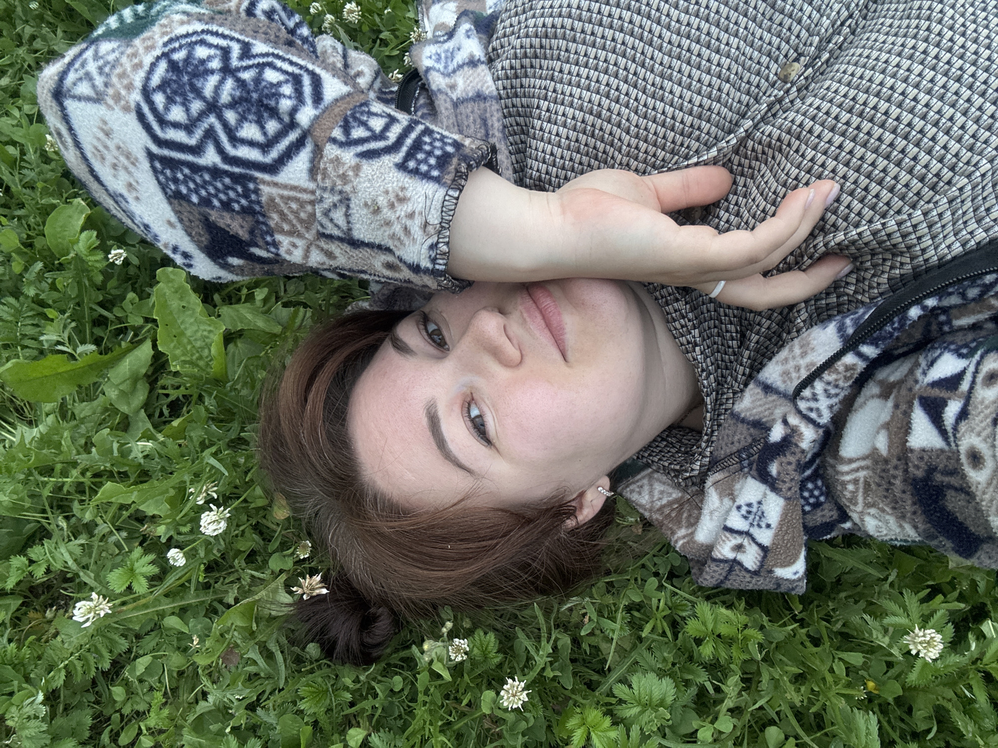 A person is lying on their back in a grassy area with small white flowers. The person has brown hair and is wearing a patterned jacket. Their right hand is raised, covering their right eye. The location is specified as Маркидоновка, Russia. The scene is outdoors.