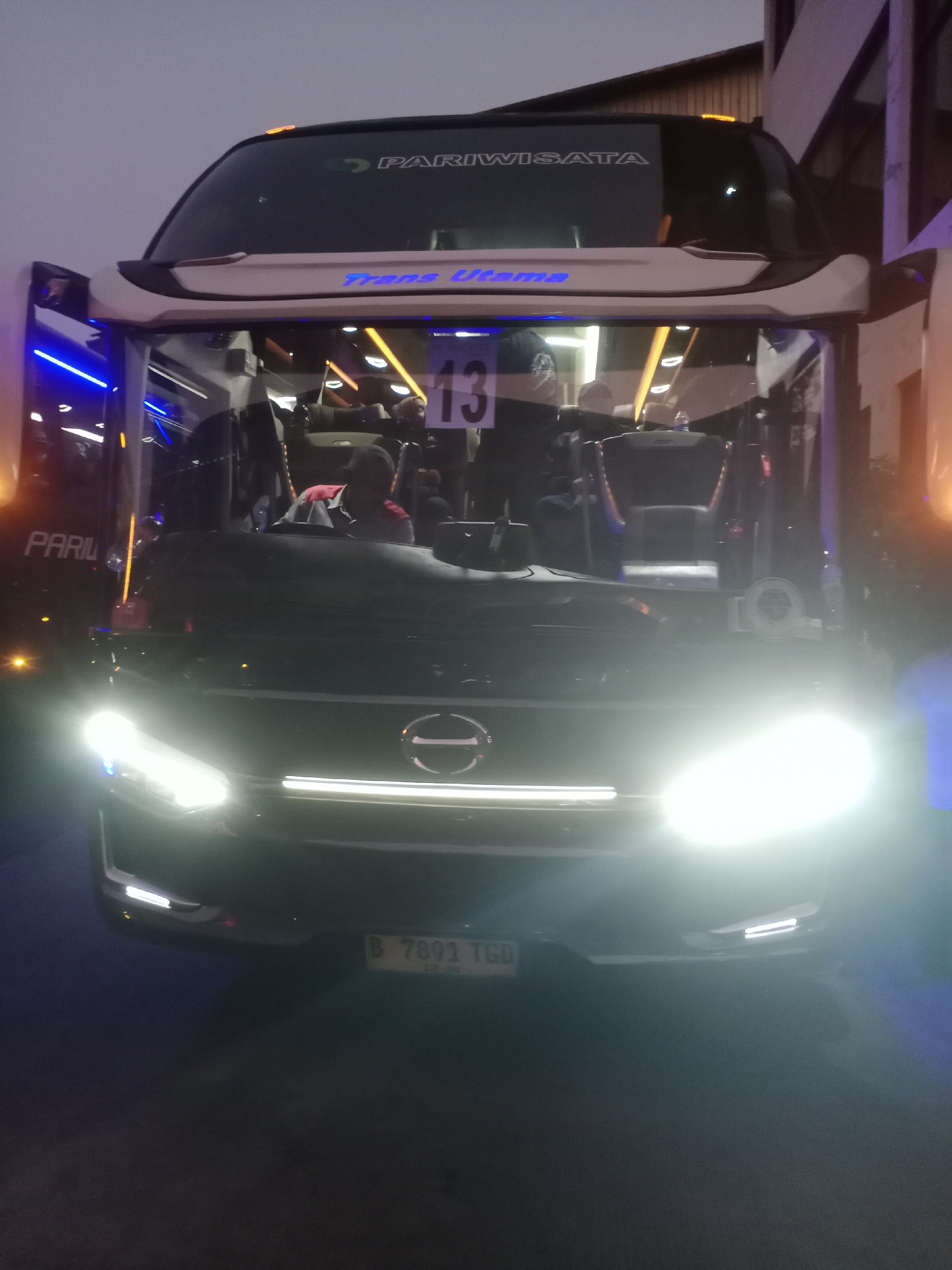 A nighttime exterior shot shows the front of a large, dark-colored tour bus with its headlights on, illuminating the scene. The bus has a white trim around its windshield and "Trans Utama" written in blue letters above the windshield. The word "PARIWISATA" is visible in white text on the upper portion of the windshield. Inside the bus, a person wearing a red shirt is visible in the driver's seat. A number "13" is displayed on the interior of the bus. A license plate with the text "B 7891 TGD" is affixed to the lower front of the bus. The scene appears to be in an urban environment, possibly a street or parking lot, with some buildings visible in the background. The sky is overcast and dark.