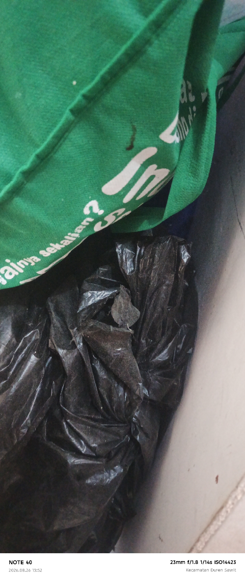 The image is a close-up shot of a green fabric item, possibly a bag or clothing, draped over a black plastic bag filled with dark material. The green fabric has white lettering on it, some of which is visible. The visible text appears to be in Indonesian, with a question mark at the end. The black plastic bag is crumpled and appears to be partially filled with debris or waste. The scene is lit from above, casting shadows on the plastic bag. The bottom of the image contains a timestamp and location information: "NOTE 40 2026.08.26 13:52" and "23mm f/1.8 1/14s ISO14423 Kecamatan Duren Sawit". This suggests the photo was taken in the Duren Sawit subdistrict of East Jakarta, Indonesia, on August 26, 2026, at 1:52 PM.