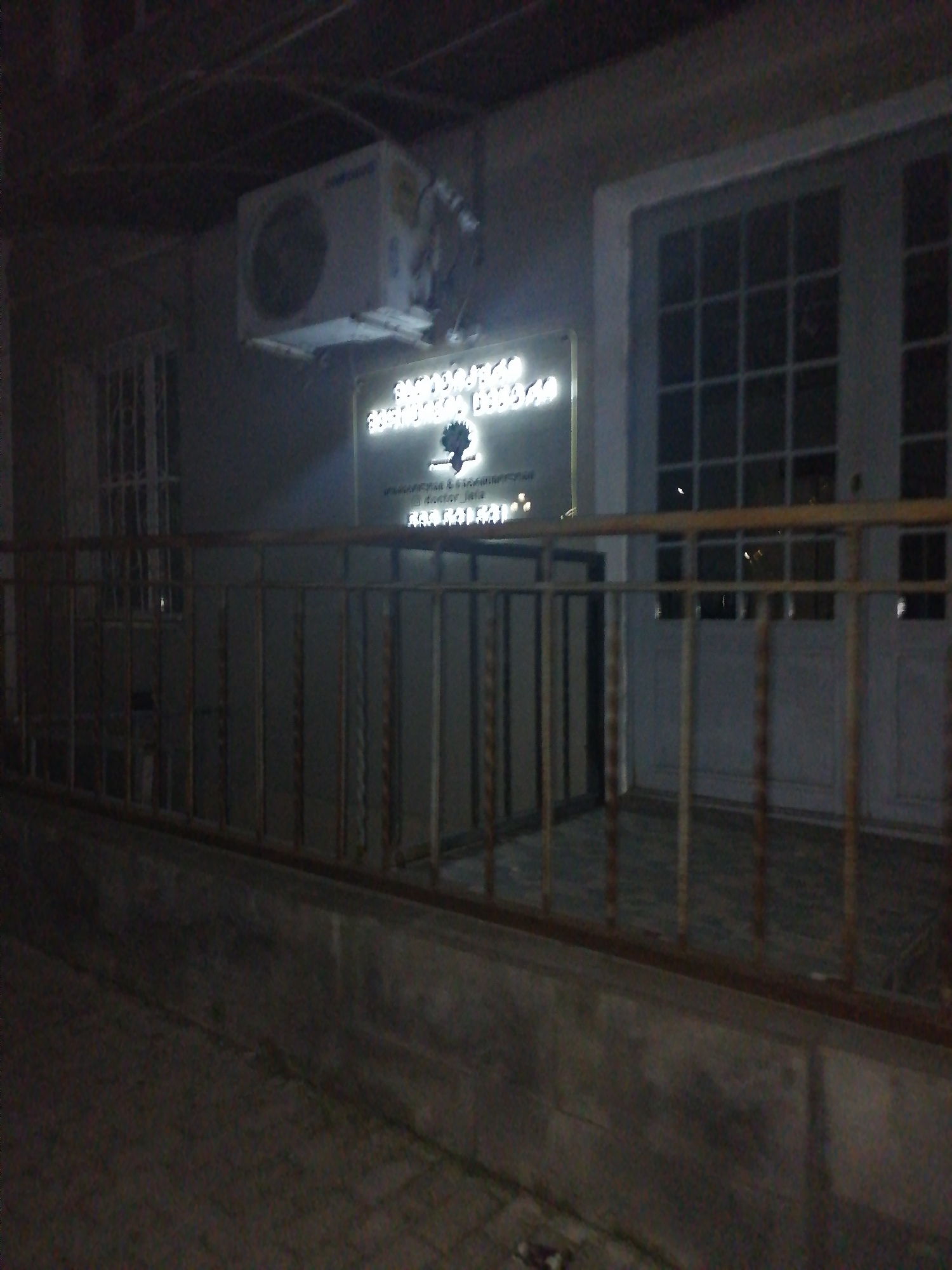 The image depicts the exterior of a building at night in Rustavi, Georgia, as suggested by the sign text, which appears to be in Georgian. The building has a light-colored facade, a large white air conditioning unit mounted on the wall, and a prominent door with multiple glass panes, possibly a double door. A metal railing runs in front of the building, obscuring some of the lower part of the structure. A sign with illuminated text and a tree logo is mounted on the building above the railing. The ground in front of the building appears to be paved. The overall lighting is dim, indicating nighttime, and the image quality is affected by low light conditions and potential motion blur.