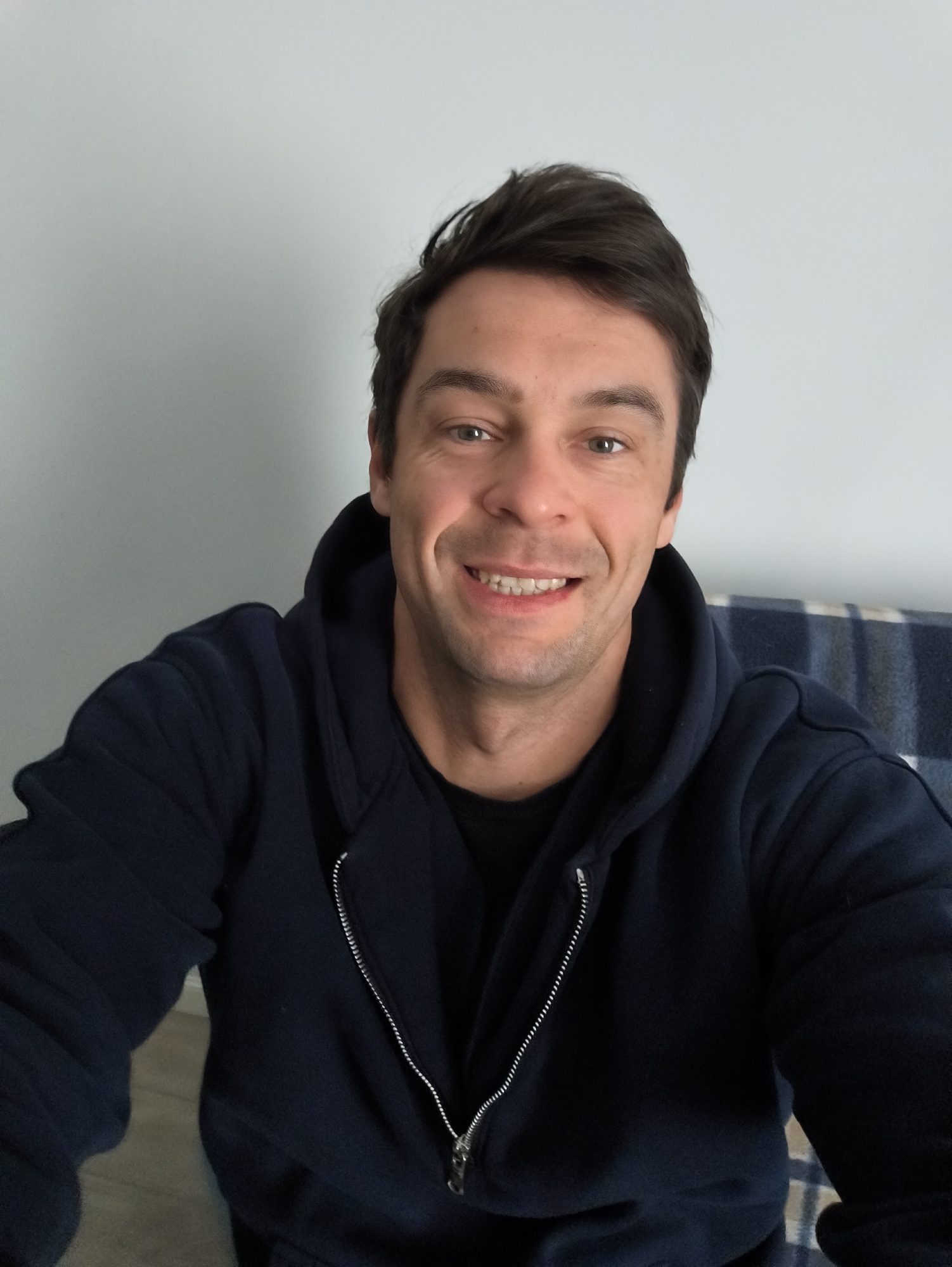 A fair-skinned man with dark hair smiles at the camera. He is wearing a dark-colored zip-up hoodie over a black shirt. The background is a plain white wall and a blue and white checkered object, possibly a blanket or part of furniture. No indication of location in Moscow, Russia.
