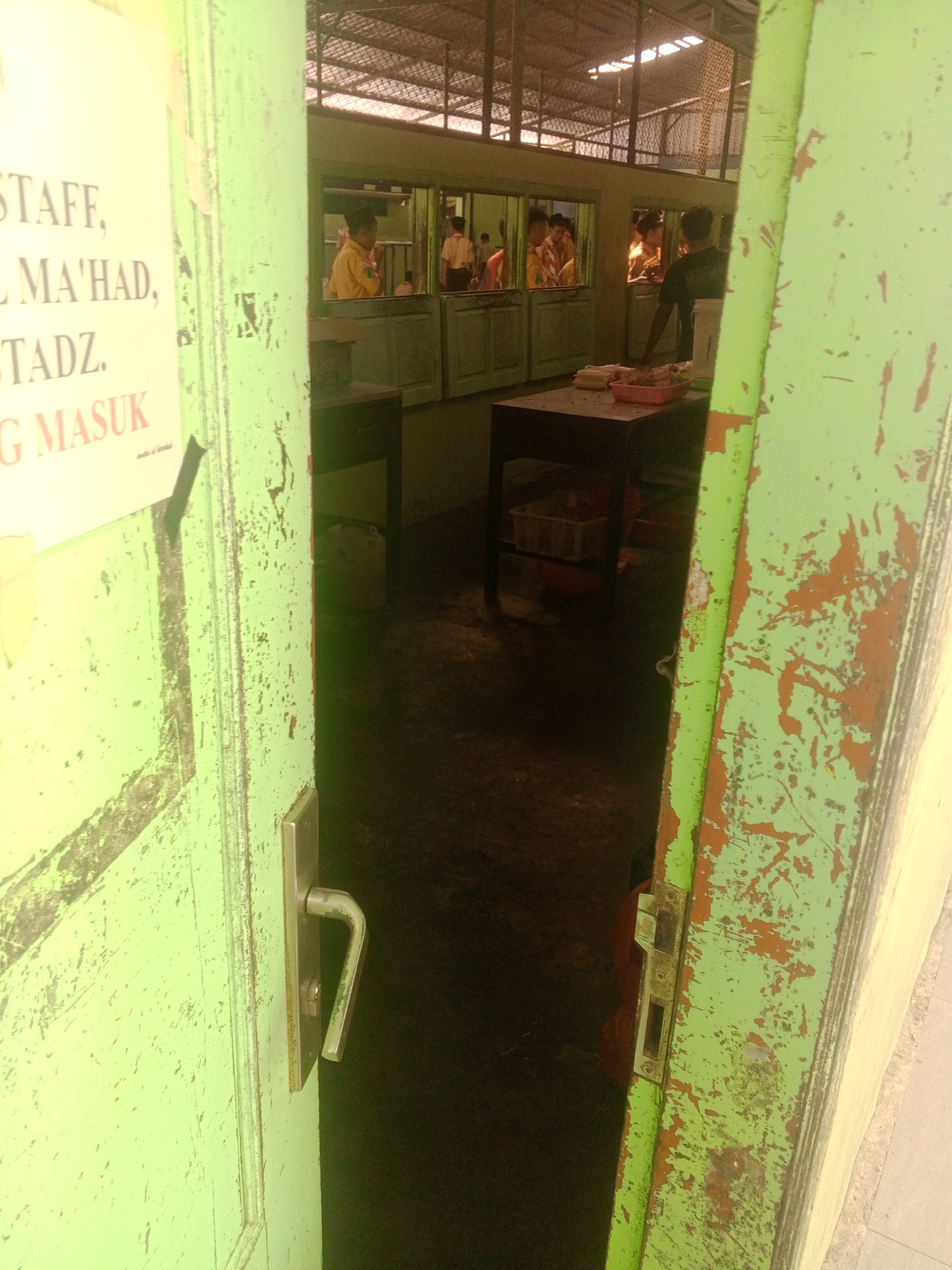 The image depicts an open doorway, revealing a dimly lit interior. The doorframe is painted a peeling light green with patches of orange visible underneath, suggesting age and wear. A metal door handle is attached to the door. To the left of the doorway, a portion of a poster is visible, featuring text in English and what appears to be Malay or Indonesian. The visible text includes "STAFF," "MA'HAD," "STADZ," and "G MASUK."
Through the doorway, the interior appears to be a room with tables and reflective surfaces, possibly windows or mirrors, showing multiple individuals. Some individuals are wearing light-colored uniforms. One person is standing behind a table laden with food items, including what looks like corn and some packaged food in a red container. The floor of the interior is dark and appears to be concrete or asphalt. The ceiling is visible in the background, with a metal grid structure.
This scene is likely located in Ponorogo, Indonesia, given the presence of text in Indonesian and the general architectural style. The environment suggests a public or institutional setting, potentially a school or a community center, given the presence of uniforms and food distribution. The overall impression is one of a casual, everyday scene within such an establishment.