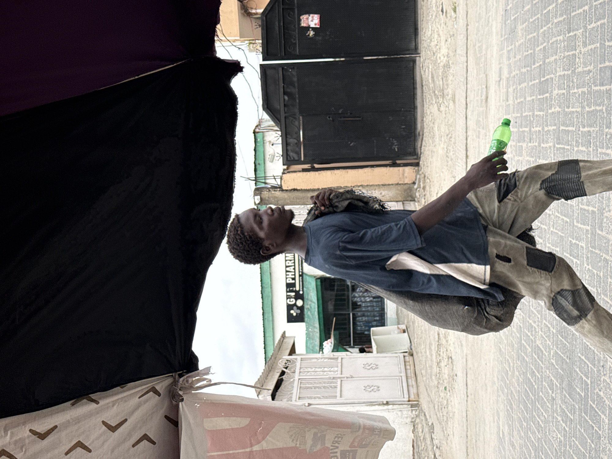 This outdoor street photograph, taken in Igboefon, Nigeria, features a young Black man captured in a candid moment. The image is rotated 90 degrees counter-clockwise, showing the man standing on a paved surface, looking upwards and to his left.
The man, who appears to be an adolescent or young adult, wears a blue short-sleeved t-shirt and light-colored pants with multiple visible patches. He holds a translucent green plastic bottle in his right hand. A dark fabric item, possibly a bag or cloth, is draped over his left shoulder. His hair is short and curly.
The ground is paved with light-colored bricks arranged in a herringbone pattern. The background reveals an urban or semi-urban environment with buildings and structures. A prominent sign for "GNB PHARM" is visible on a building with a green awning, indicating a pharmacy. Large, dark metal gates are also visible further in the background. The left side of the foreground is occupied by dark fabric, possibly part of a tent or market stall, with a section of white fabric displaying brown chevron-like patterns beneath it.
The scene appears to be set during the daytime, with soft, diffused lighting. The man's expression is neutral and observant as he gazes upwards. The image depicts an everyday street scene.