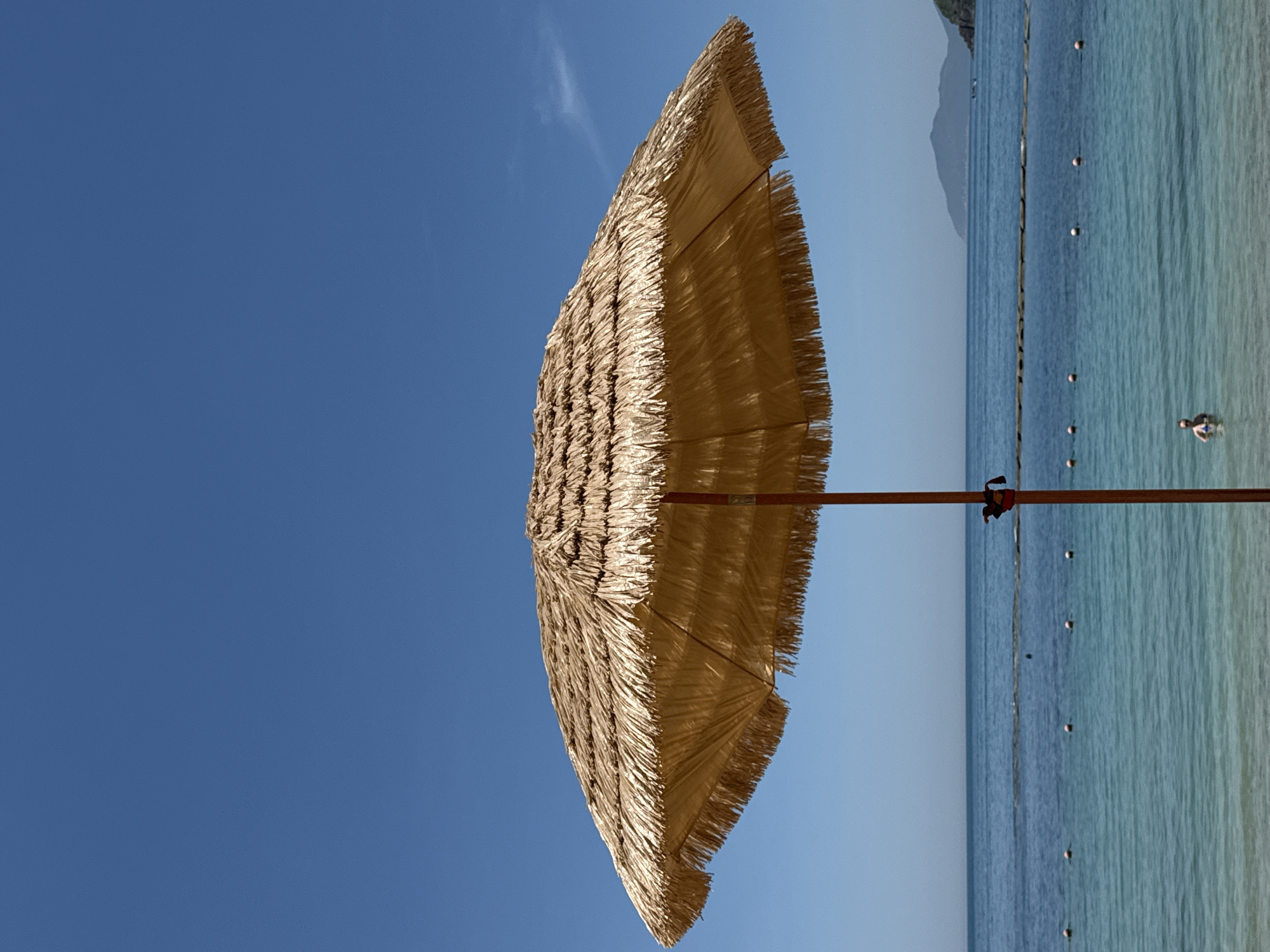 This media file shows a tilted view of a thatched beach umbrella against a clear blue sky. The umbrella is the primary object, angled towards the viewer, its straw texture and frayed edges clearly visible. To the right of the umbrella, the scene transitions to a calm, blue ocean. The water is clear, with a few white buoys marking a boundary. In the distance, faint mountains can be seen under a hazy sky. A single person is visible in the water, partially submerged, appearing to be swimming or wading. The overall setting is a tropical beach. The image appears to have been taken during the daytime, under sunny weather conditions, with the clear sky indicating good weather. The orientation of the image is unusual, suggesting it was shot with the camera held vertically and then rotated. No text is visible in the media file.