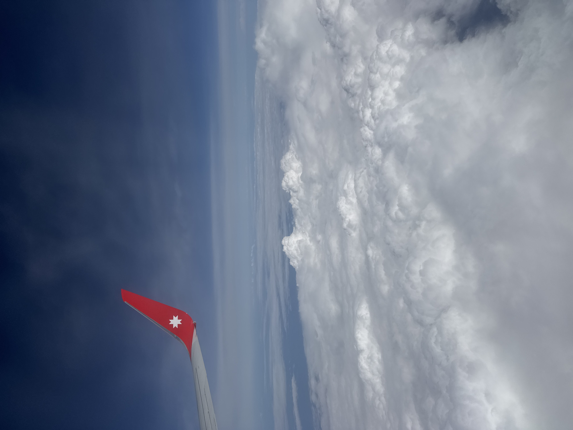 The image shows an airplane wing in flight, with a red wingtip bearing a white star. The sky is visible, with a clear blue expanse transitioning into a band of layered clouds. The clouds are textured and appear dense, filling much of the view. The image appears to have been taken from inside the airplane, looking out at the wing and the sky. The location in the city of Lyamtsino, Russia is not apparent from the provided details.