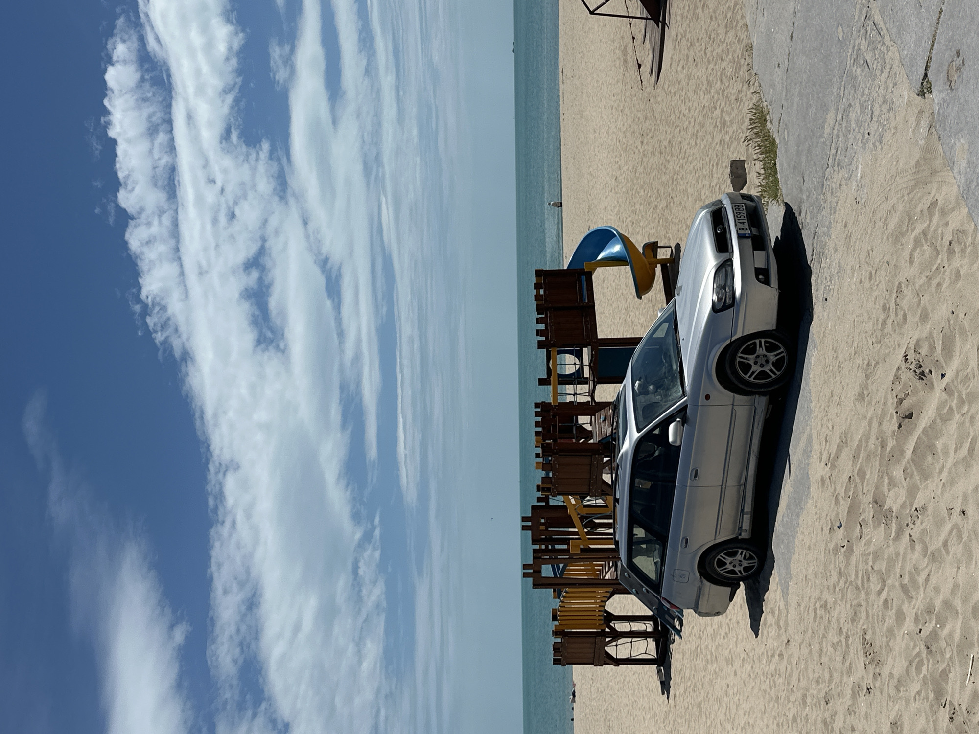 A silver car with a Bulgarian license plate is parked on a sandy beach next to a wooden structure, likely a covered seating area. The Black Sea and a cloudy sky are visible in the background. The location is likely a beach area in Varna, Bulgaria. There are no visible people in the immediate vicinity.