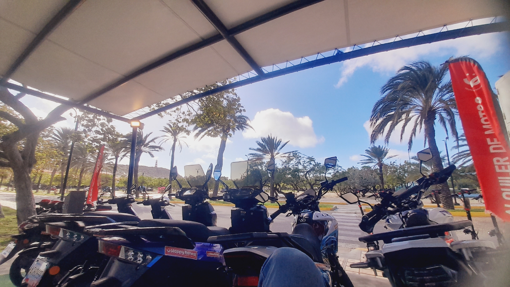 The image depicts a row of parked scooters and motorcycles under a covered structure with a white canopy and dark metal beams. In the background, palm trees and other vegetation are visible, along with a lighthouse on a hill. The sky is blue with scattered clouds. A red banner with white lettering, possibly advertising scooter rentals, is displayed prominently to the right. The scene appears to be an outdoor rental area, possibly in a coastal or tropical location. While the location is stated to be Pedro González, Venezuela, this cannot be definitively confirmed from the visual information alone. No people are clearly visible engaging in any actions, although a person's leg in jeans is visible in the foreground, suggesting someone may be seated on or near one of the scooters. The lighting suggests it is daytime.