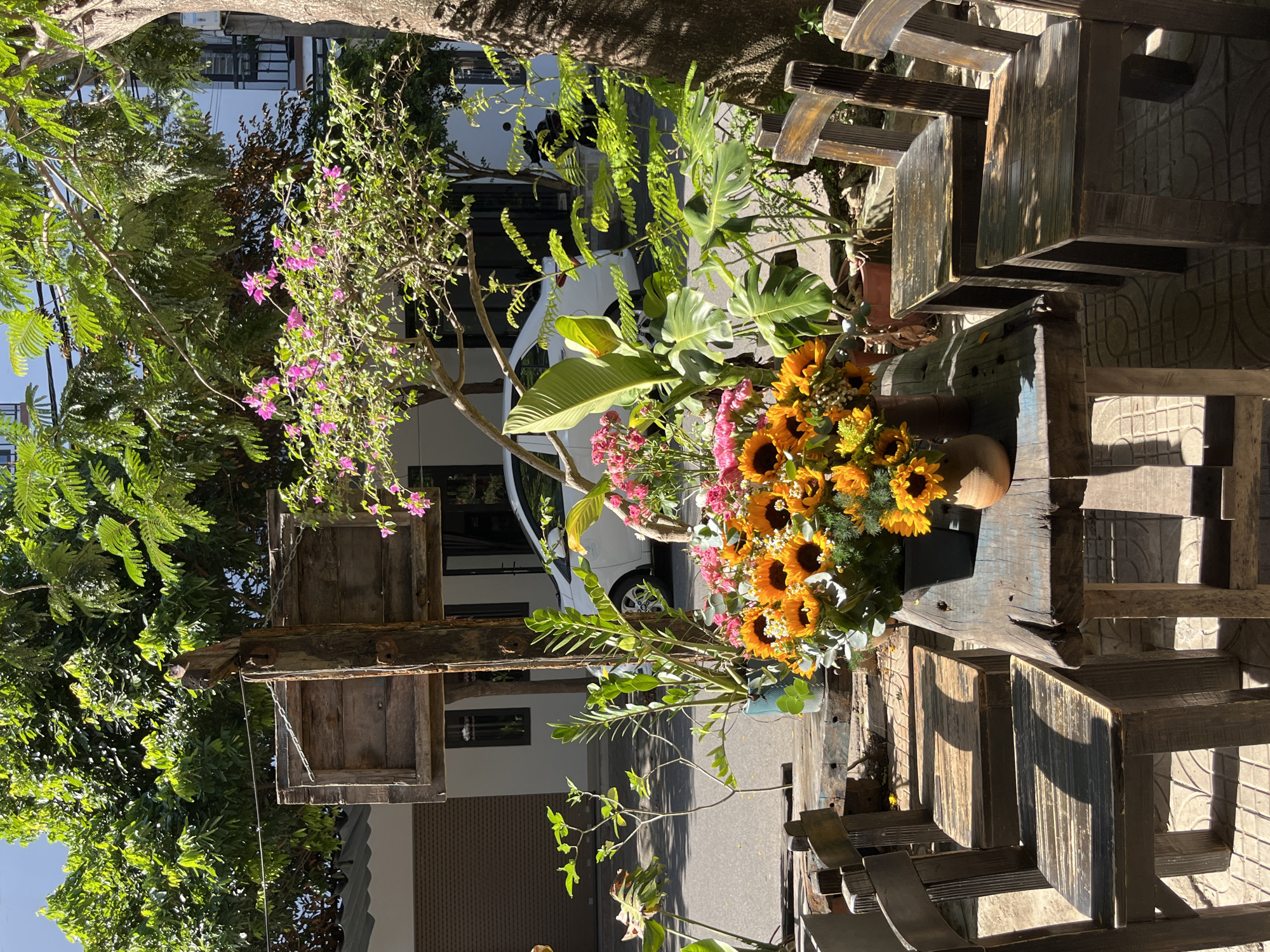 The image depicts an outdoor seating area in front of a building, likely a cafe or restaurant, on a sunny day in Đà Nẵng, Vietnam. In the foreground, a weathered wooden table is set with two vases of artificial flowers, one with sunflowers and the other with pink and yellow blooms. Four dark wooden chairs are arranged around the table. Behind the table, a white car is parked on the street. Lush green trees and plants surround the seating area, with vibrant pink bougainvillea flowers blooming on a bush. The building in the background has a white facade and appears to be a residential or commercial structure. The overall atmosphere is relaxed and inviting, suggesting a peaceful spot to enjoy the outdoors.
