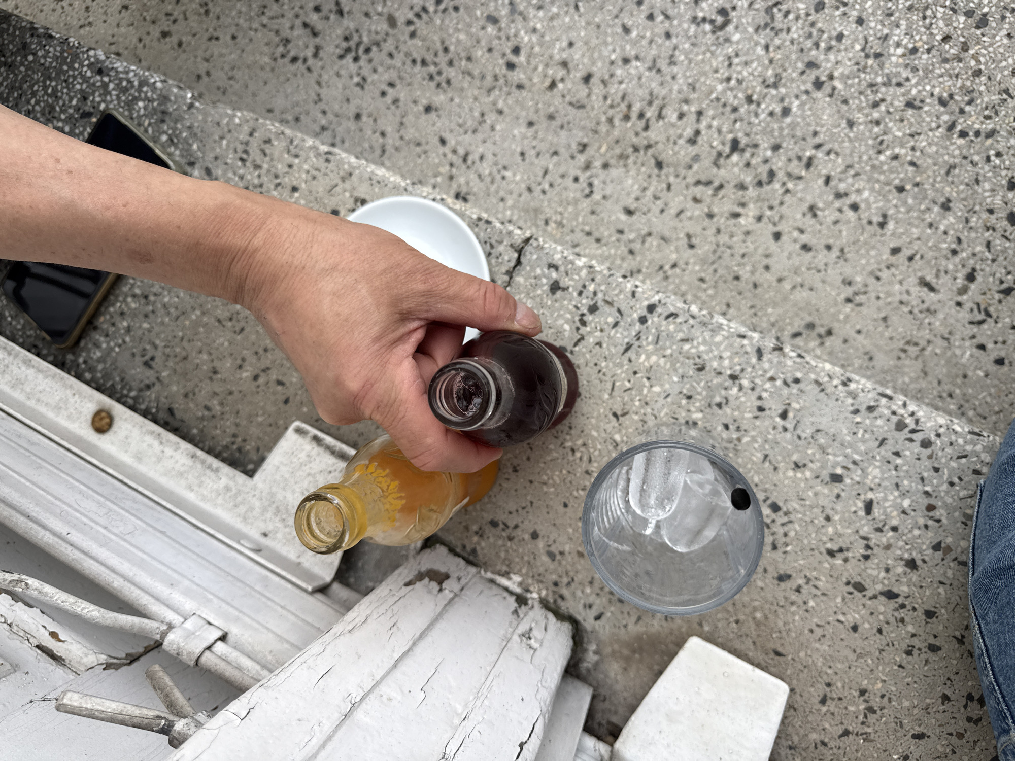 In Sofia, Bulgaria, a hand is reaching for a small, dark glass bottle of liquid, seemingly to pour it into a clear glass filled with ice. Next to this bottle, a yellow glass bottle of orange liquid stands upright. A white bowl is visible behind the bottles. The scene is set on a textured, light-colored concrete surface, possibly a sidewalk or outdoor seating area, with a white painted wooden structure in the background. A portion of a mobile phone is visible on the concrete surface. The lower part of the image shows the top of a person's blue jeans.