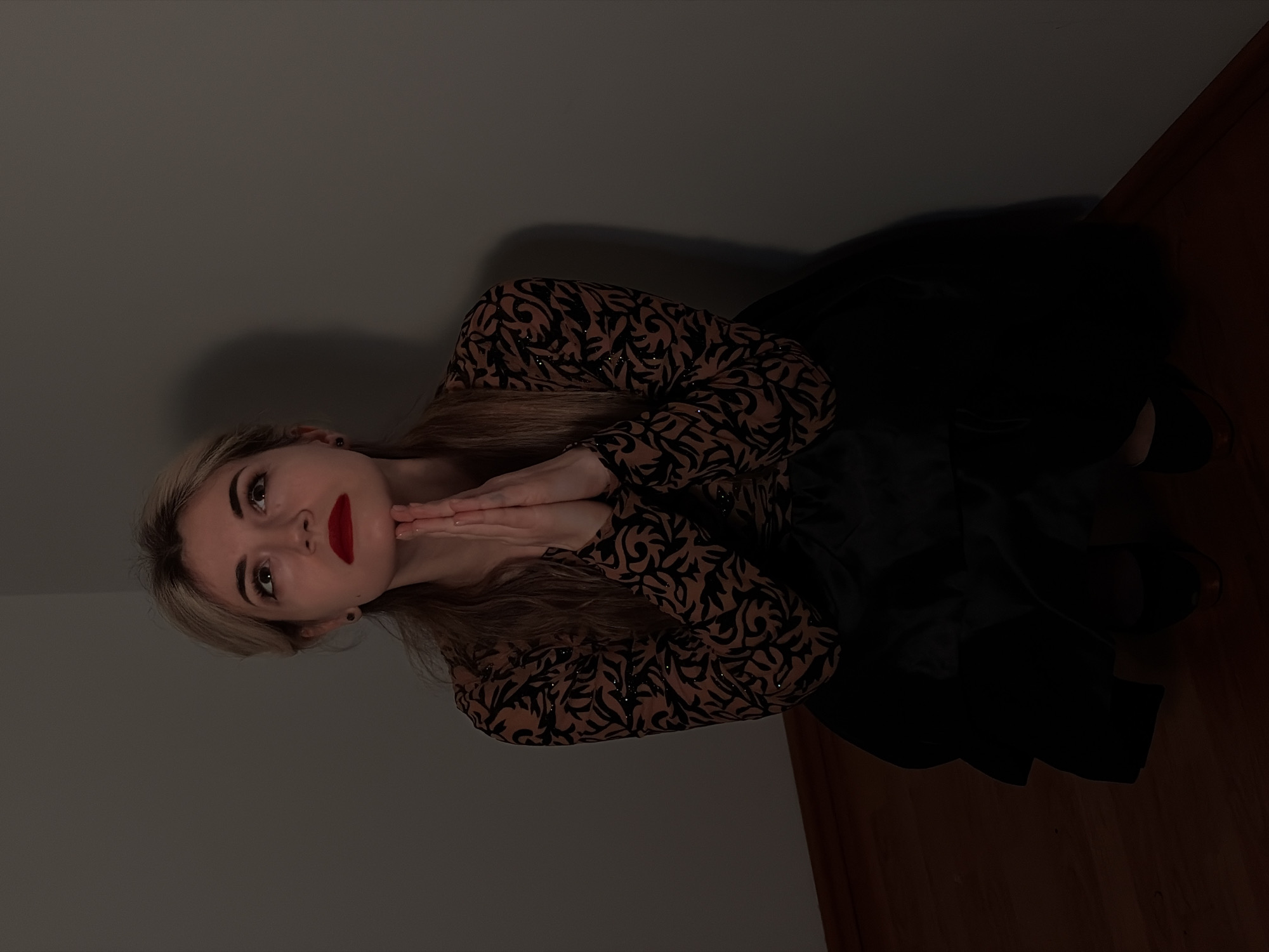 A female individual is depicted kneeling or seated on a wooden floor within the corner of a room. She has blonde hair, dark eyes, and is wearing red lipstick. Her attire includes a long-sleeved top featuring a brown and black intricate pattern with subtle shimmering accents, and a dark, voluminous skirt. Her hands are pressed together in front of her chest, fingers pointing upwards, in a supplicant gesture. Her head is tilted slightly, and her gaze is directed upward. The environment consists of plain, light gray walls converging at a corner, and a reddish-brown wooden plank floor with a dark baseboard. The scene is dimly lit, with light primarily illuminating the subject's upper body from the left. No specific details are present within the image to determine a precise location in Budva, Montenegro.
