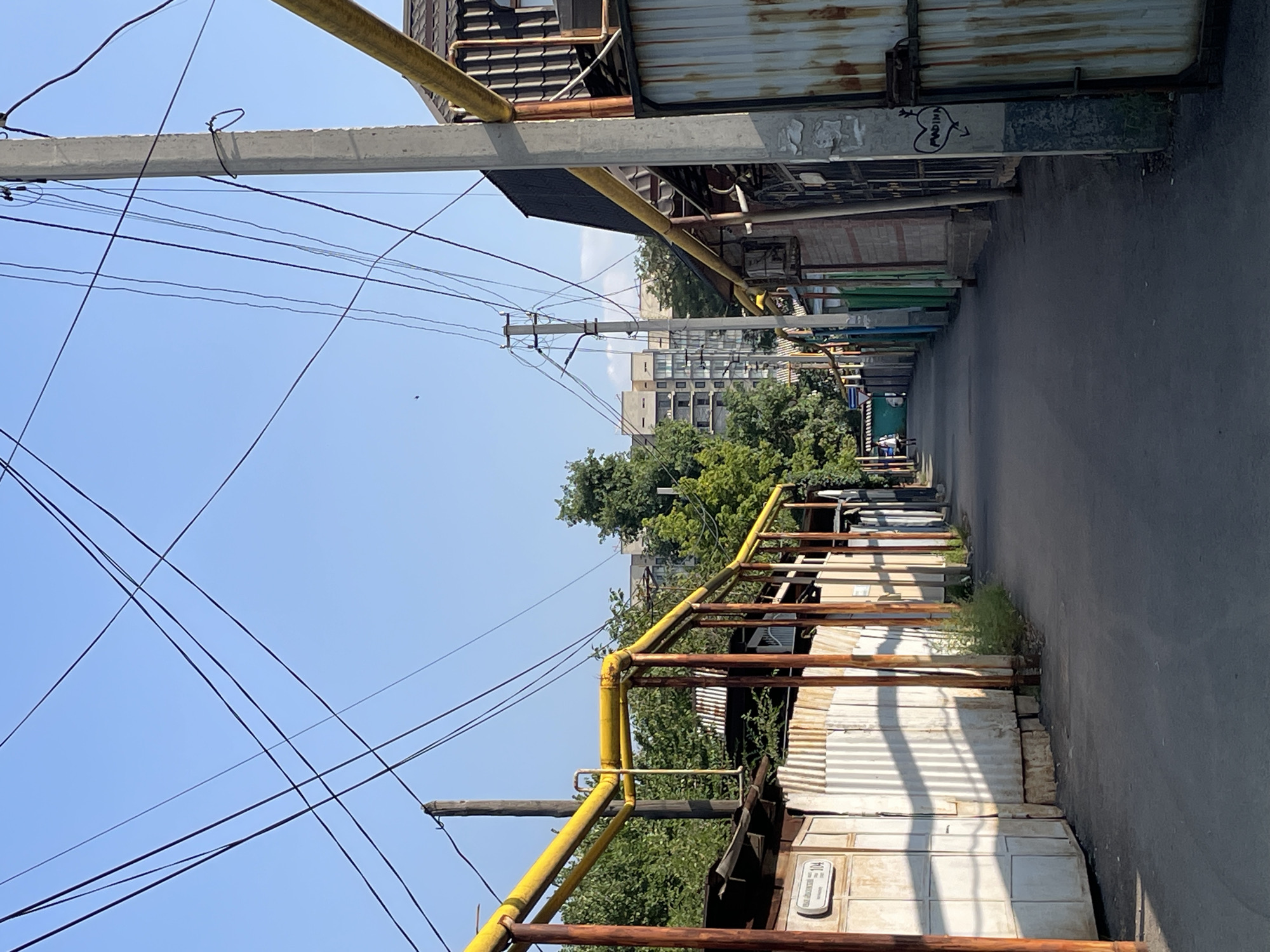 The image depicts a narrow, paved alleyway in Almaty, Kazakhstan, lined with low-rise structures and overhead utility lines. The alley is flanked on the left by a corrugated metal fence with a visible street sign indicating "ул. Айтеке би" and the number "104". A bright yellow pipeline runs along the top of this fence, supported by rusty orange metal poles. On the right, a similar corrugated metal fence is present, with graffiti of a heart and an arrow on a concrete pole. Further down the alley, a single person is walking away from the camera. In the background, taller, modern apartment buildings are visible behind a line of trees. The sky is clear and blue, suggesting a sunny day. The scene appears to be a residential or service alleyway within the city.