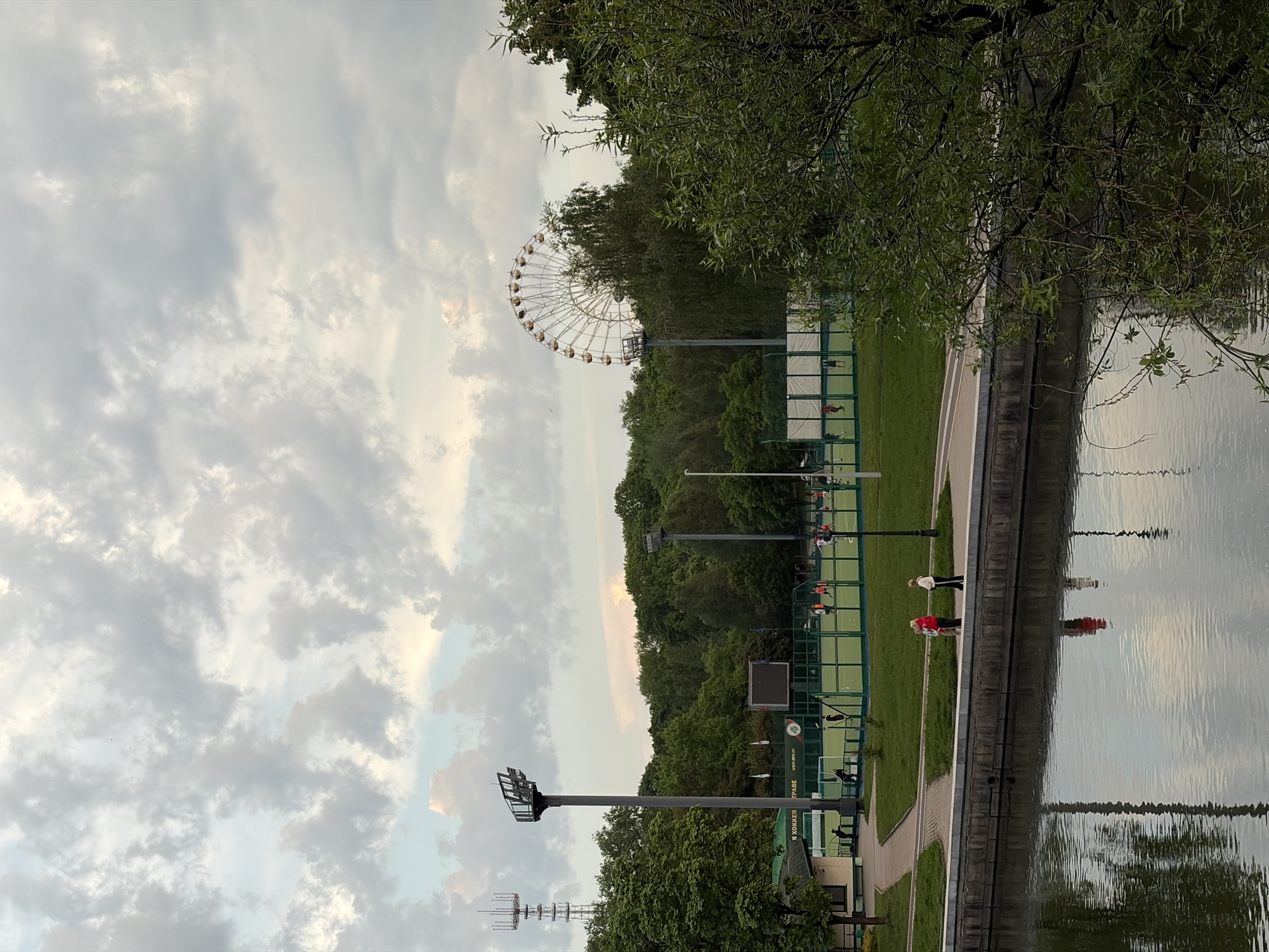 This photograph shows a park in Minsk, Belarus. In the background is a ferris wheel, partially obscured by trees. In the foreground there is a body of water. A path runs between the water and a green space with tennis courts. Several people are visible along the path and on the courts. Above, the sky is filled with clouds. There are light posts along the path and surrounding the tennis courts.