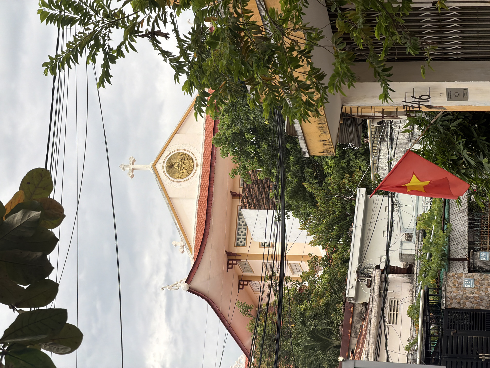 The image depicts a street scene in Nha Trang, Vietnam. In the background, a light pink building with a red tiled roof, characteristic of Vietnamese architecture, is visible. The building has decorative elements on its facade, including a circular medallion with a golden relief and a cross at its peak, suggesting it is a church or religious institution. In the foreground, a Vietnamese flag with a yellow star on a red background is prominently displayed. Overhanging the scene are power lines and lush green foliage from trees, indicating an urban environment. The sky is overcast, suggesting a cloudy day. The presence of residential buildings and gates in the lower part of the image further defines the urban setting.