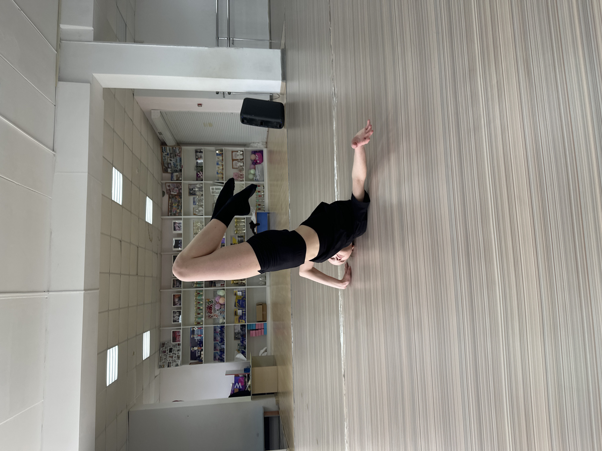 A person in black athletic wear is on a light-colored hardwood floor with vertical stripes. The person's feet are flexed in black socks. One of the person's arms is stretched towards the side. In the background, there is a wall with fluorescent lighting and a grid of pictures on a shelf. Also visible is a black box mounted on the wall. The location is a dance or exercise studio. I do not have information to confirm the location in the city of Votkinsk, Russia.