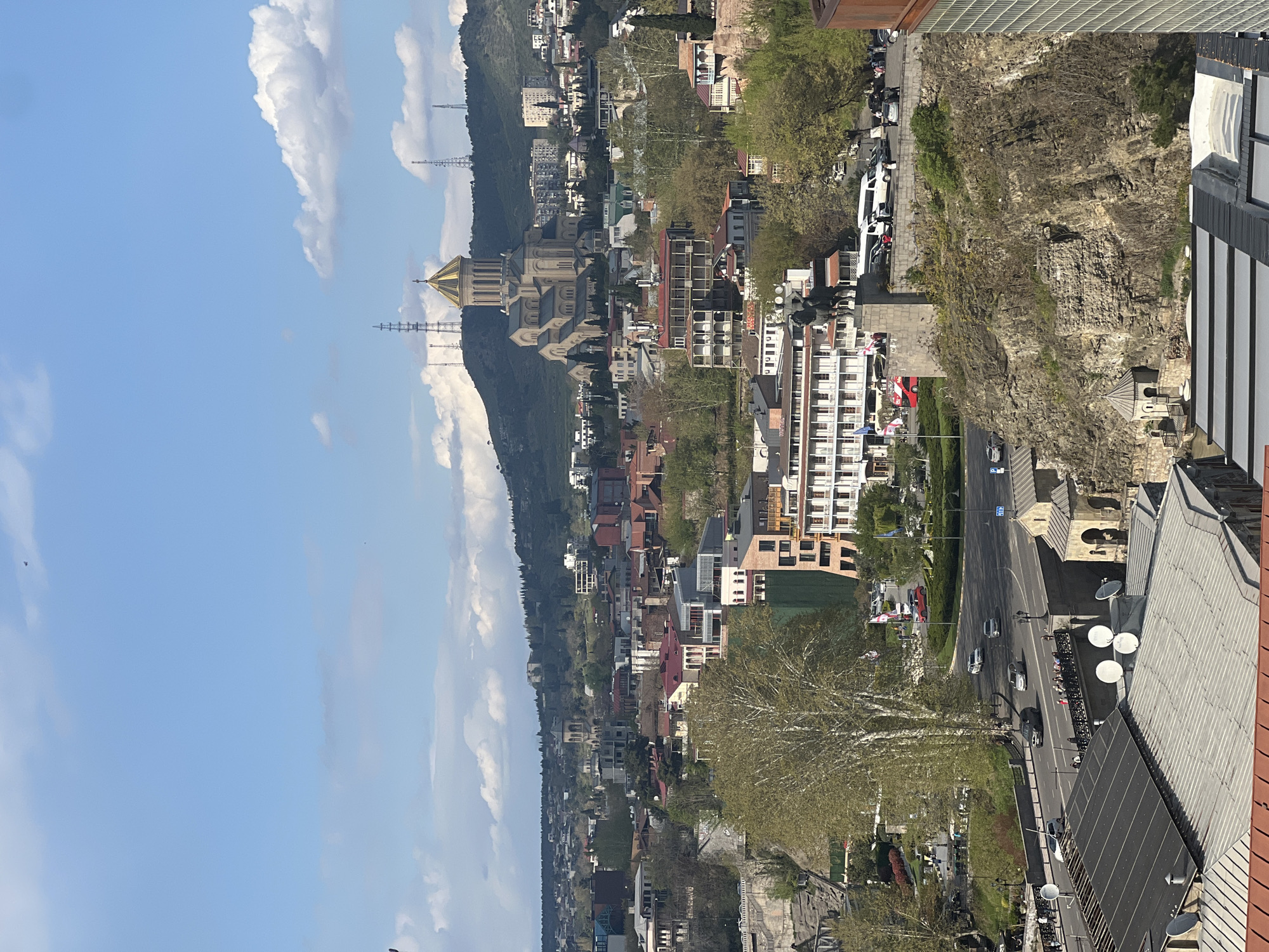 This image captures a panoramic, elevated view of the city of Tbilisi, Georgia, bathed in bright daylight under a clear blue sky dotted with fluffy white clouds. The cityscape unfolds across hilly terrain, with numerous buildings, both residential and commercial, sprawling from the foreground into the distant hills.
A prominent landmark, the Holy Trinity Cathedral of Tbilisi (Sameba Cathedral), with its distinctive golden dome and light stone facade, stands majestically on a hilltop in the mid-ground, surrounded by other structures. The buildings exhibit a mix of architectural styles, including modern multi-story blocks and older buildings with red and grey tiled roofs. Many rooftops feature white satellite dishes.
In the foreground and mid-ground, a multi-lane road winds through the urban landscape, carrying several vehicles including cars and at least one white bus. Pedestrians can be seen on sidewalks. To the right, a steep, rocky cliff face rises, showing exposed rock strata with sparse vegetation. Green trees with fresh, light foliage are scattered throughout the city, indicating spring or early summer.
Distant hills, covered in dense greenery and featuring what appear to be telecommunication towers, frame the horizon. The bright sunlight suggests a clear, pleasant day, likely morning or early afternoon, creating clear visibility and soft shadows. The overall scene depicts a vibrant and expansive urban environment. A "P" for parking is barely discernible on a sign near the road.