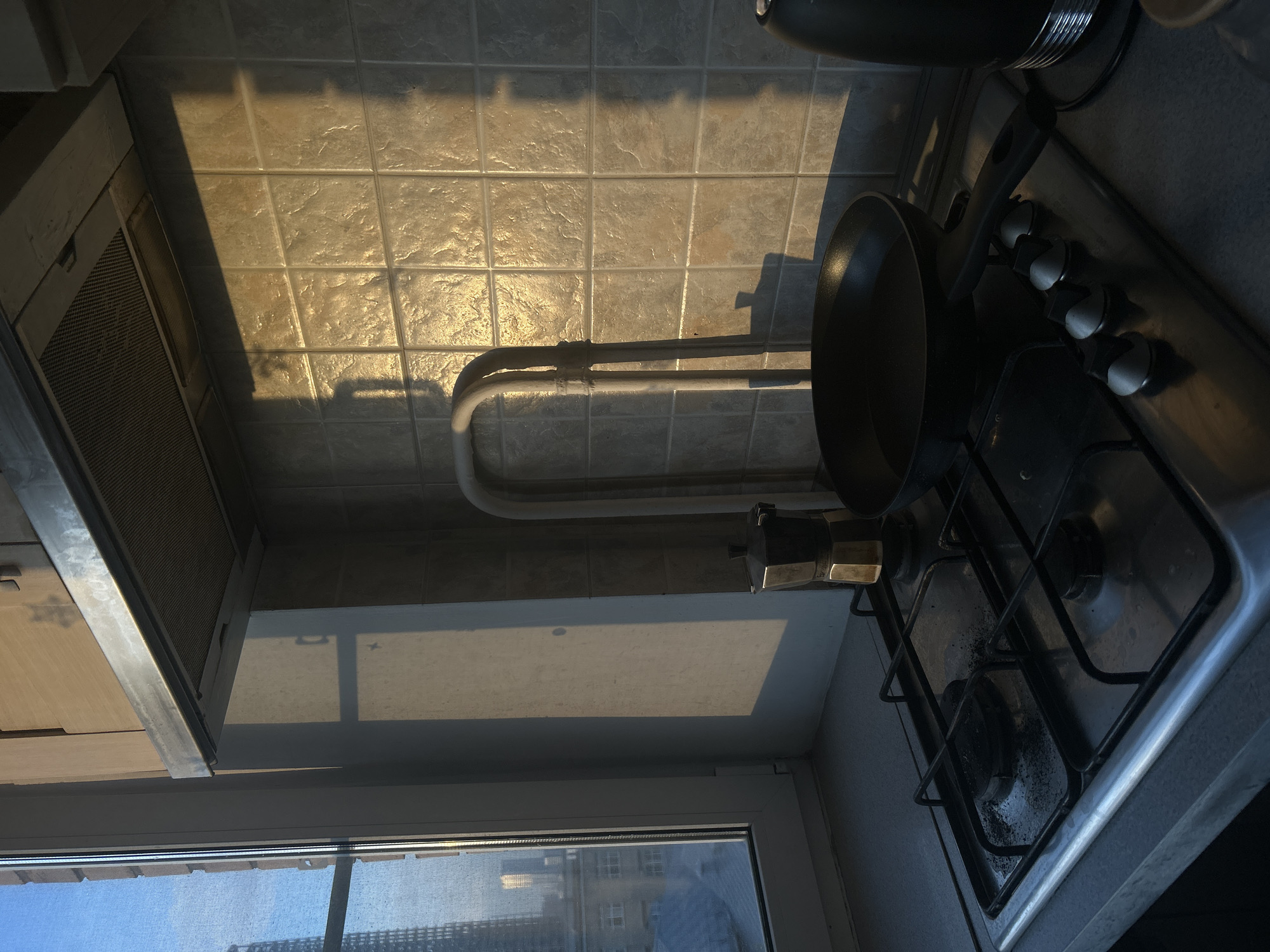 The image depicts a sunlit kitchen corner, likely in an apartment setting. No people are visible. The main subjects are a gas hob, a frying pan, a moka pot, an extractor hood, and a tiled wall.
On the right side of the frame, a gas hob with two visible burners and grates is situated on a dark countertop. A black frying pan rests on the hob's grates, with its handle pointing towards the viewer. A silver moka pot is positioned behind the frying pan. The hob features several control knobs along its front edge.
Above the countertop, a wall is covered with light-colored, square tiles that have a textured surface. Two white pipes run horizontally across this tiled wall, located behind the hob. On the left side of the image, an extractor hood with a metallic frame and mesh filter is mounted on the wall.
The scene is illuminated by natural light, with golden sunlight streaming in from a window on the lower left. This light casts long, distinct shadows of the pipes, the extractor hood, and other kitchen elements onto the tiled wall and countertop, indicating either early morning or late afternoon. The sun glints off the textured tiles, creating bright highlights.
Through the window, a clear sky is visible, along with glimpses of multi-story buildings, suggesting an urban or suburban environment. The window itself has a white frame and appears to have a screen or fine netting.
The overall impression is a quiet, domestic scene bathed in warm sunlight. There is no visible activity taking place. No discernible text appears in the image. Given the location context of Khimki, Russia, the architectural style visible through the window and the kitchen setup are consistent with apartment living in the region.