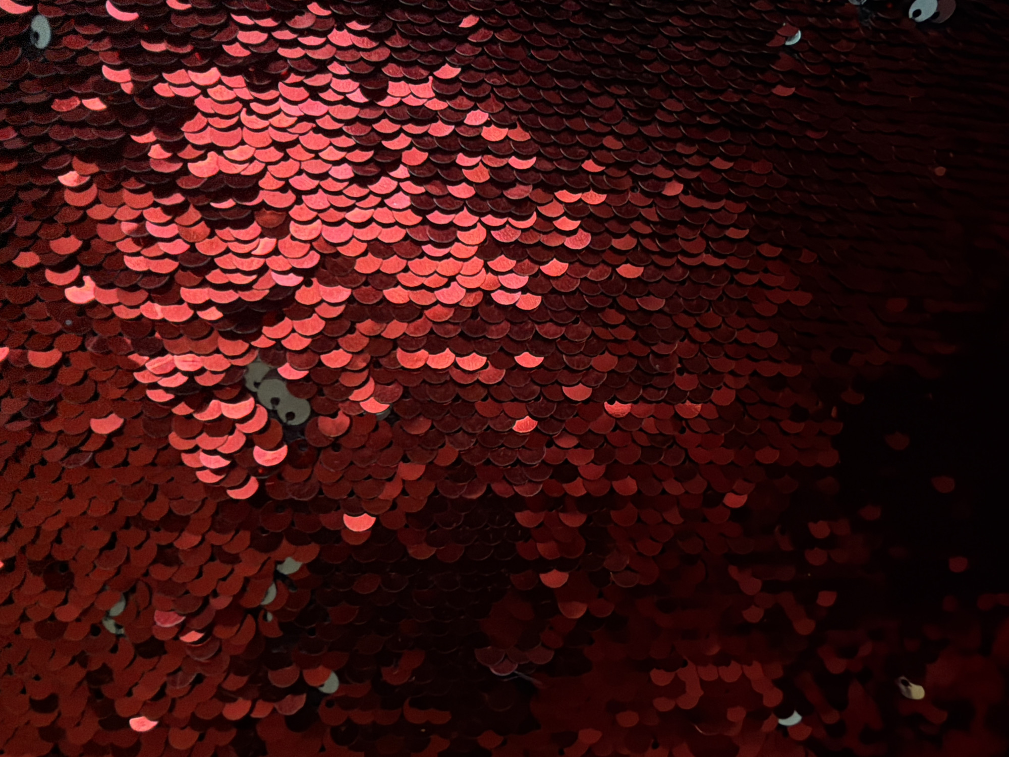 The image is a close-up, overhead shot of a surface covered in small, circular, red sequins. The sequins are densely packed and overlap each other, creating a textured, shimmering effect. The lighting casts some shadows and highlights, giving the sequins a three-dimensional appearance. The background is dark and out of focus, drawing all attention to the sequined surface. The color palette is dominated by various shades of red, with some lighter, almost white highlights where the light catches the sequins. There are no people, objects, or discernible environmental features within the frame, suggesting this is an abstract detail shot of a material.