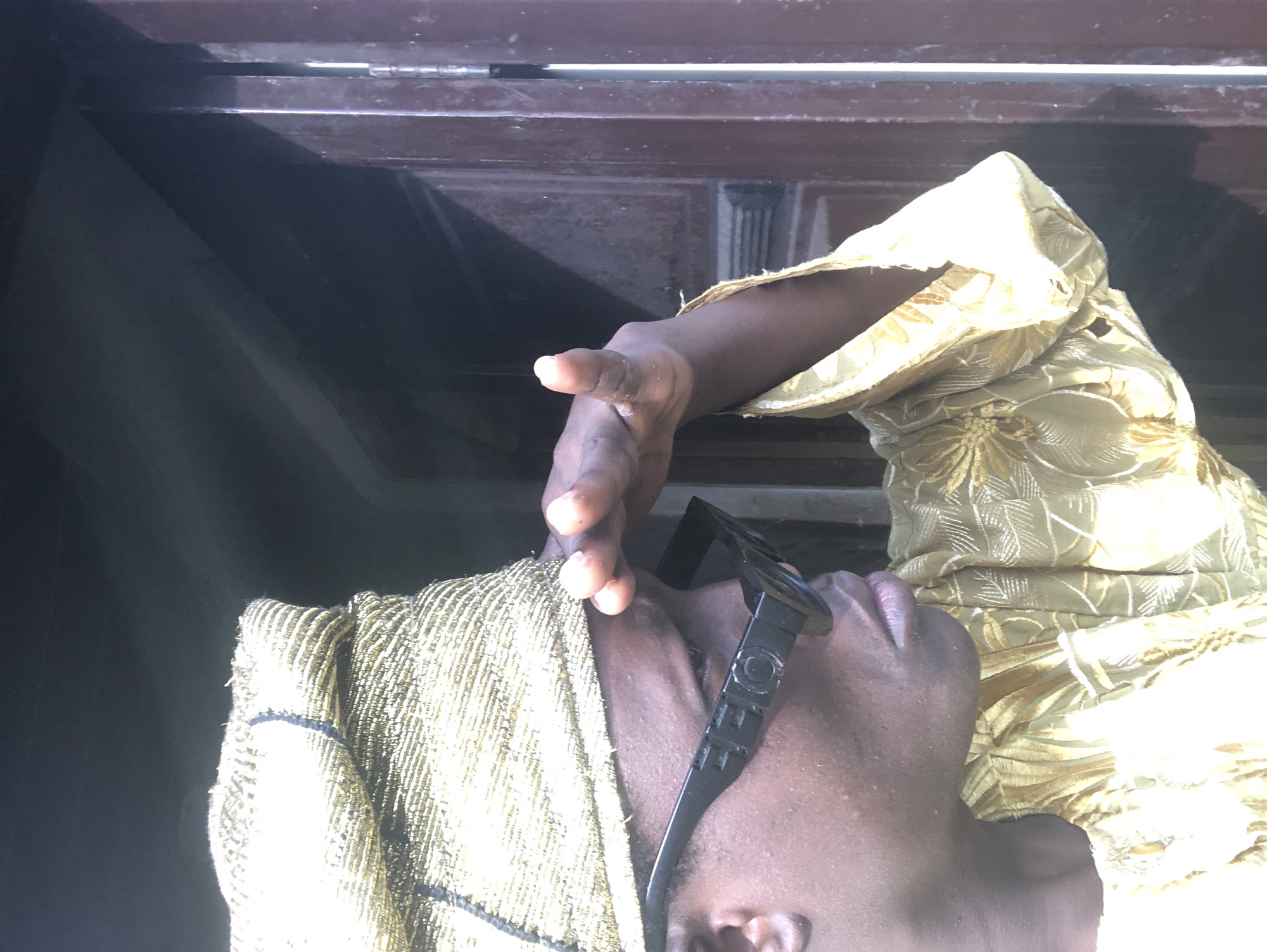 The image shows a person wearing a gold beaded cap and a gold embroidered outfit. The person is also wearing black sunglasses. Their right hand is raised, shielding their eyes from the sun. The background appears to be a dark doorway or opening, with a brown wooden door frame visible on the right. The scene is outdoors, with sunlight illuminating the person and their clothing. The image's composition suggests the person is looking out from an interior space into a brighter area. The location is Ado, Nigeria.