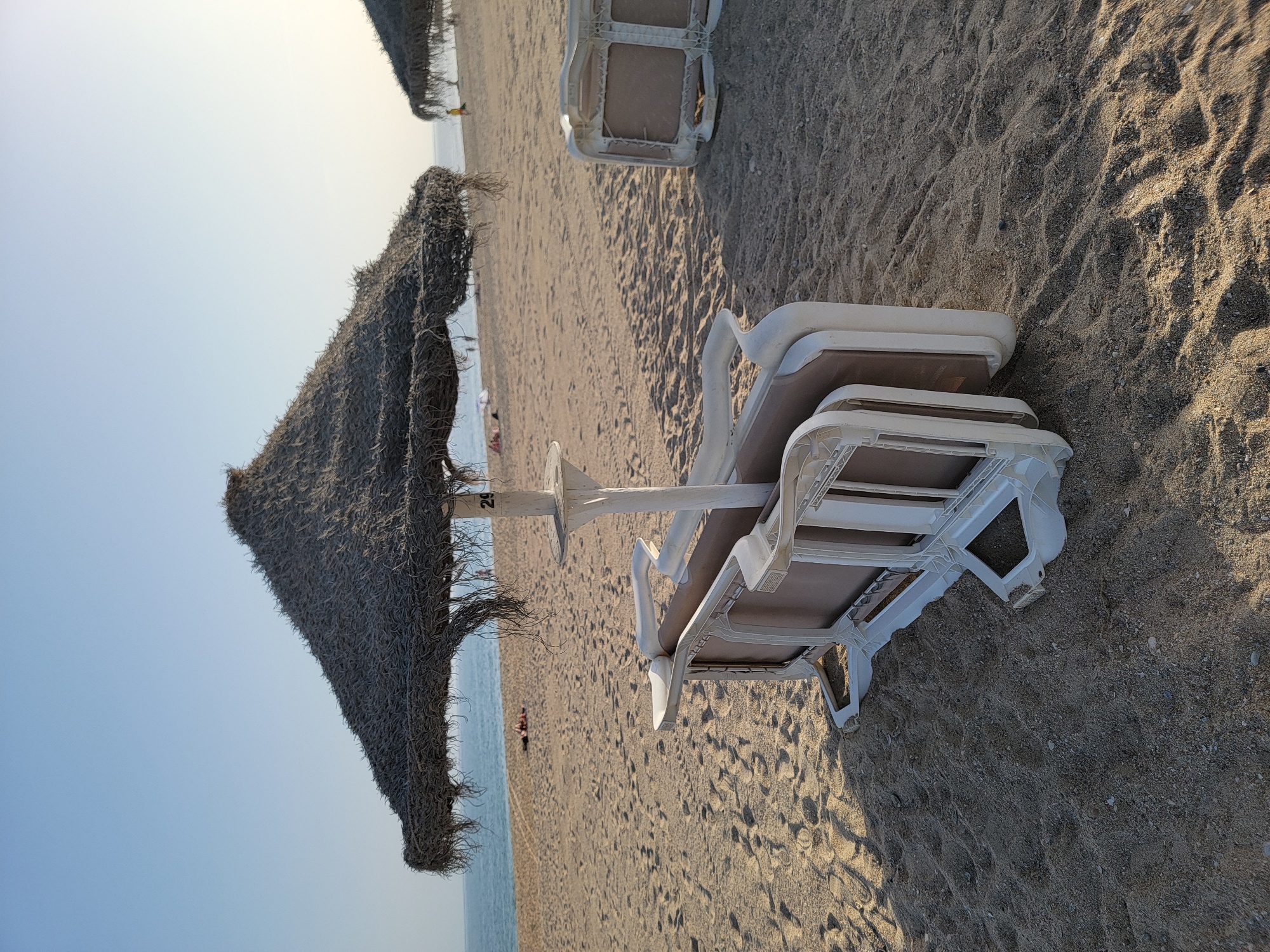 The image depicts a sandy beach in Quarteira, Portugal, with a thatched umbrella and stacked lounge chairs in the foreground. The umbrella, made of dry palm fronds, provides shade over the chairs, one of which has the number '29' visible on its pole. The chairs are white plastic with beige fabric seating, stacked on top of each other and slightly askew. The sand is a light brown color, with small footprints visible. In the background, the ocean meets the horizon under a clear, pale blue sky. Other thatched umbrellas and a few distant figures can be seen on the beach. The overall scene is tranquil, suggesting a relaxed atmosphere on a sunny day.