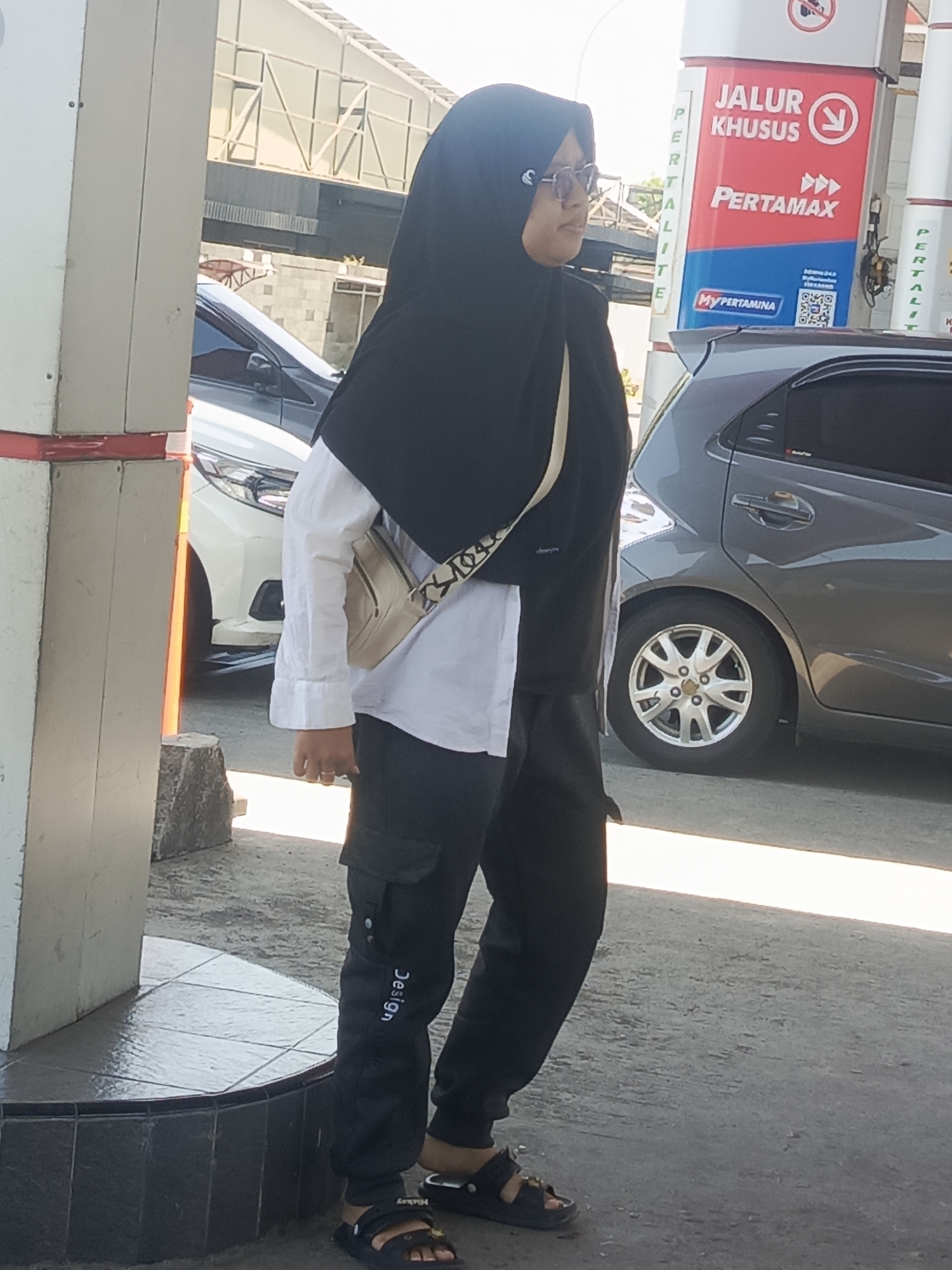 A medium shot captures a Muslim woman wearing a black hijab, glasses, and a white long-sleeved shirt layered under a black vest. She is also wearing black cargo pants and black sandals. A beige crossbody bag is slung over her shoulder. She is standing next to a white and red gas pump at a Pertamina gas station in Waru, Indonesia. Behind her, a grey car is parked, and a white car is visible in the background. The scene appears to be outdoors during the daytime, with a paved surface for vehicles and a tiled platform around the gas pump. A sign on the gas pump reads "JALUR KHUSUS PERTAMAX" and "MyPertamina". Another sign visible on the pump has the text "PERTALITE" repeated vertically.
