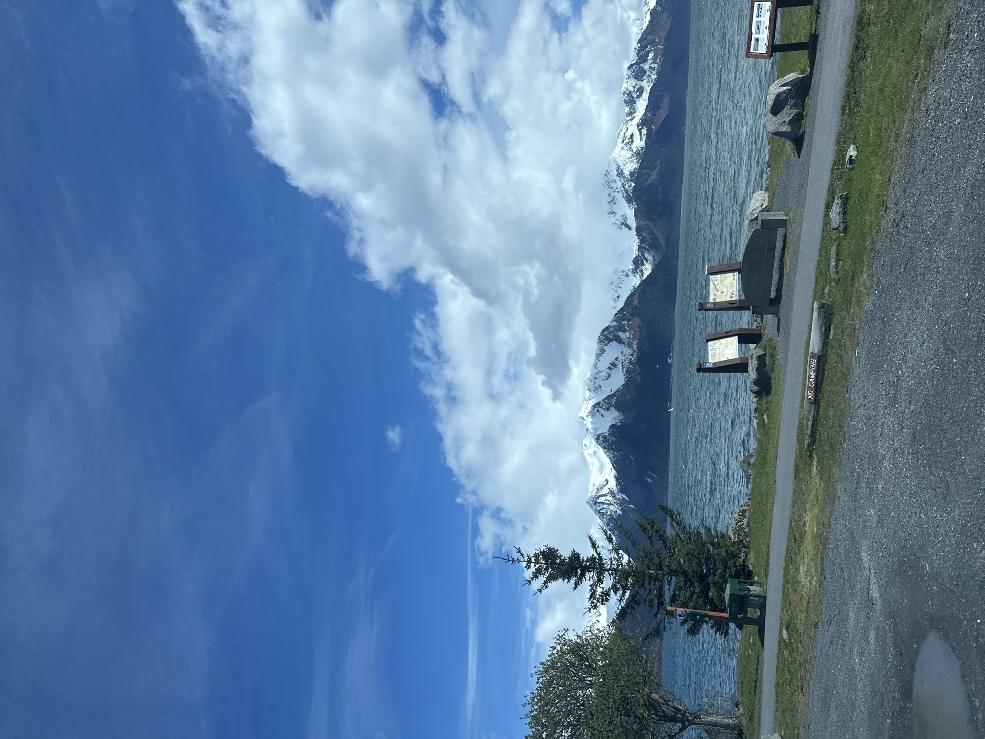 The image presents a landscape view likely taken in Seward, Alaska, United States of America. In the background, snow-capped mountains form a scenic backdrop alongside a body of water, possibly a lake or a fjord. The sky is predominantly blue with patches of white, puffy clouds. The middle ground features greenery, including trees. In the foreground, there's a paved area or path, likely for pedestrian use. A "NO CAMPING" sign is visible. There are also wooden information boards placed along the path.