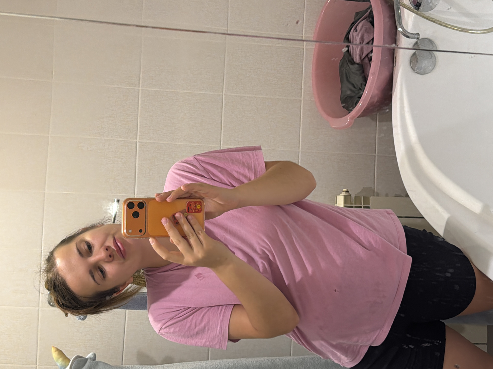 An indoor mirror selfie captures a young woman in a bathroom, likely in Sochi, Russia. The woman, with light skin and brown hair pulled back, is seen smiling gently towards the camera. She is wearing a pink short-sleeved t-shirt and black shorts, and holds an orange-cased smartphone with both hands to take the picture.
The bathroom features light beige or cream-colored tiled walls. A white bathtub is prominently visible on the right side of the frame, with chrome plumbing fixtures including a faucet and shower hose. Beside the bathtub, a pink plastic basin or bucket is placed on the floor, containing dark-colored clothes. A small portion of what appears to be a plush toy, possibly a unicorn given the visible horn and ear, is reflected in the mirror at the bottom left of the image. The overall setting suggests a casual, everyday moment. No discernible text is visible on the phone case or elsewhere in the reflection.