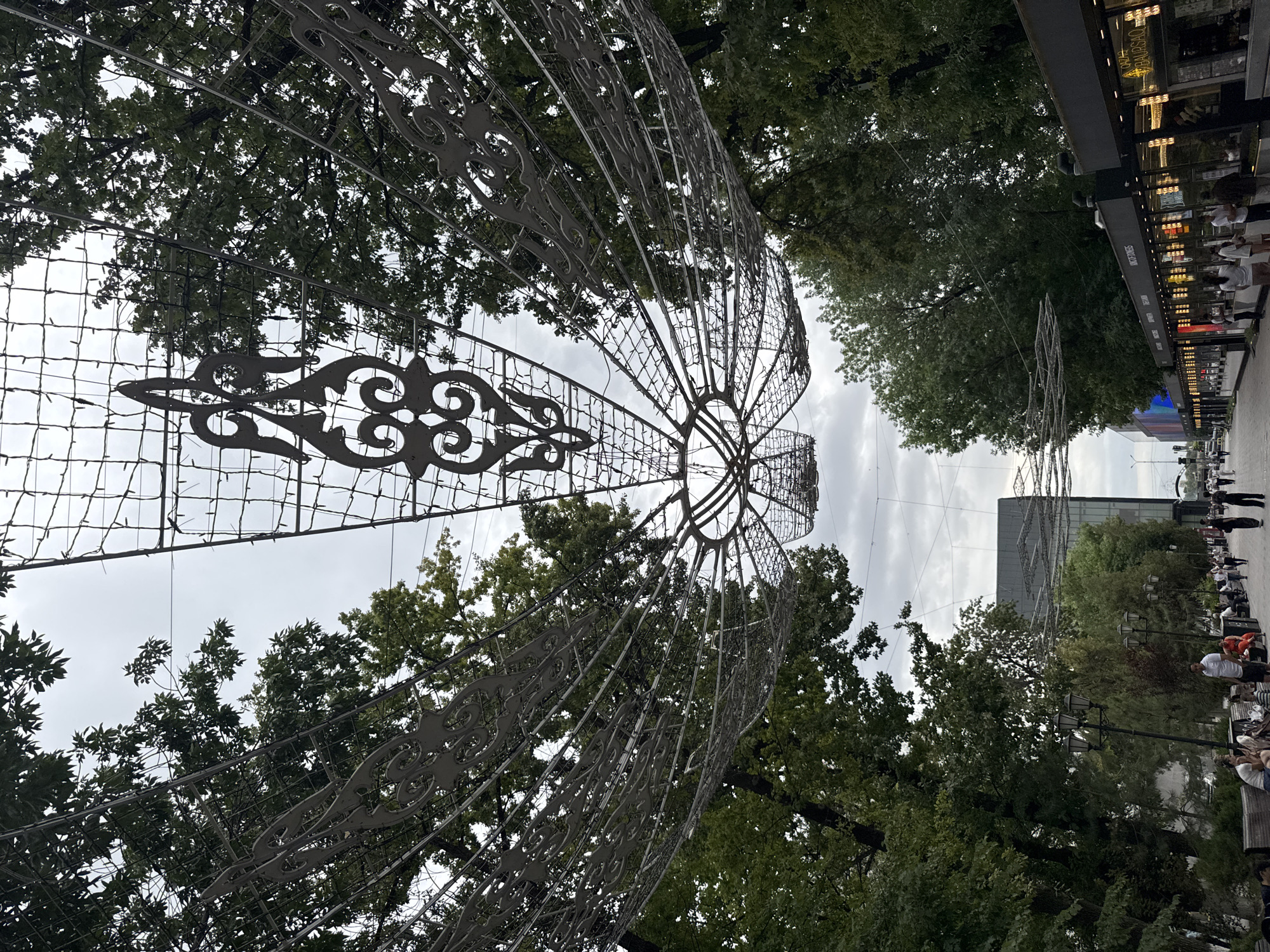 In Shymkent, Kazakhstan, an overhead decorative metal structure, resembling a fan or a large stylized flower with intricate scrollwork and lit by numerous small lights, spans across a pedestrian walkway lined with mature trees on both sides. The sky is overcast, suggesting a cloudy day. Below the trees, a pathway is visible with benches occupied by several people. On the right side of the pathway, a row of shops or food stalls with illuminated windows faces the walkway, and more people are seen standing and walking along this area. The architectural style of the shops appears modern. The overall scene depicts a public urban space with decorative elements, pedestrian activity, and commercial establishments.