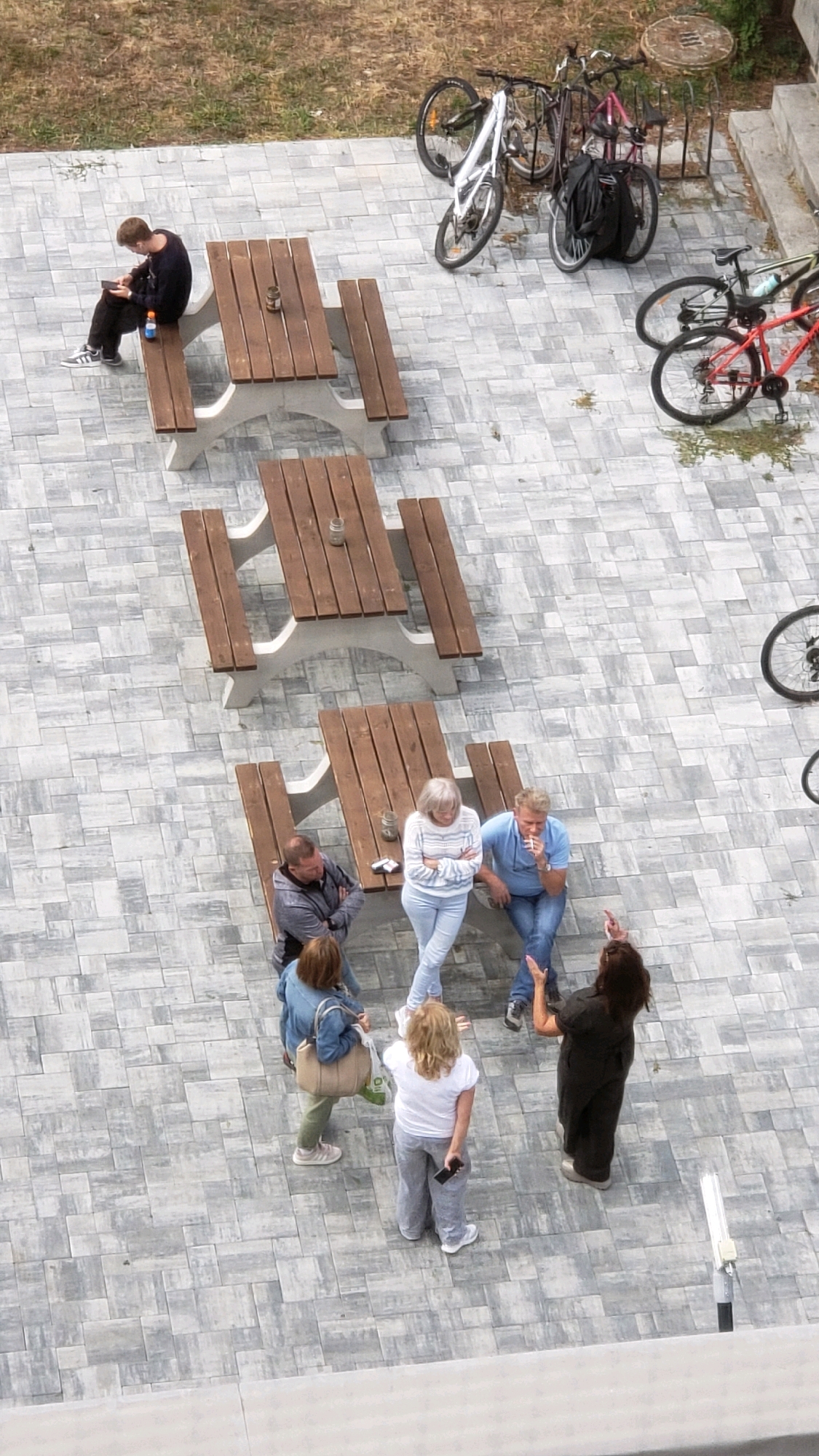 An aerial view captures a paved outdoor seating area with several picnic tables. In the background, multiple bicycles are parked. In the foreground, a group of people are gathered around a picnic table. One person sits alone at a separate table, looking at a phone. The environment suggests a public space, possibly a park or a cafe patio. The exact location in Prešov, Slovakia is not determinable from the image.