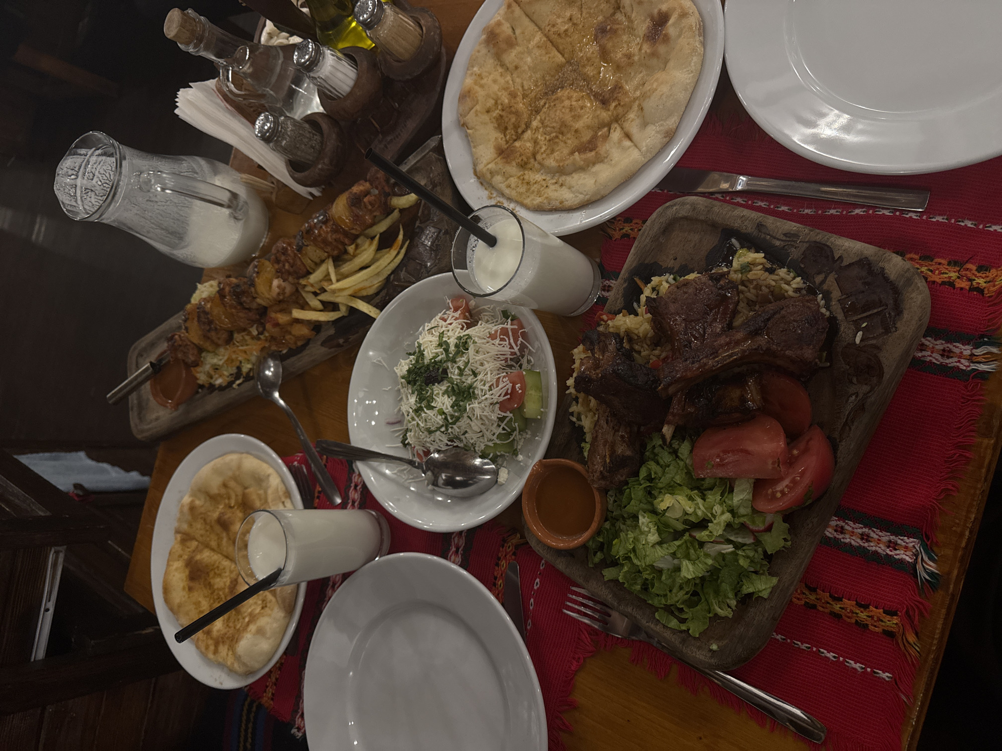 A high-angle shot captures a wooden table laden with Bulgarian cuisine, potentially in Sofia, Bulgaria. A red cloth with decorative fringes lies on the lower right corner. The food items include a large flatbread, salad with shredded cheese, grilled meat with rice on a wooden platter, and skewers of meat with fries. Glasses of what appear to be ayran (a yogurt drink) with black straws are also visible. There are also bottles of oil and vinegar in a wooden holder, salt and pepper shakers. Plates and utensils are scattered around the various dishes. The wooden table is set in what appears to be an indoor dining setting.