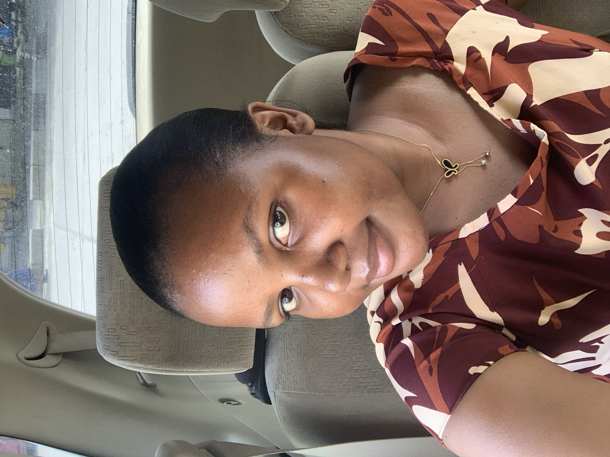 A woman is taking a selfie while seated in a car, likely in Dar es-Salaam, Tanzania. The woman is of African descent, with short black hair and brown skin. She is wearing a brown and beige patterned blouse and a gold necklace with a butterfly pendant. The car's interior is beige. The background outside the car window is blurry, with vertical white lines and a hint of greenery, typical of a cityscape.