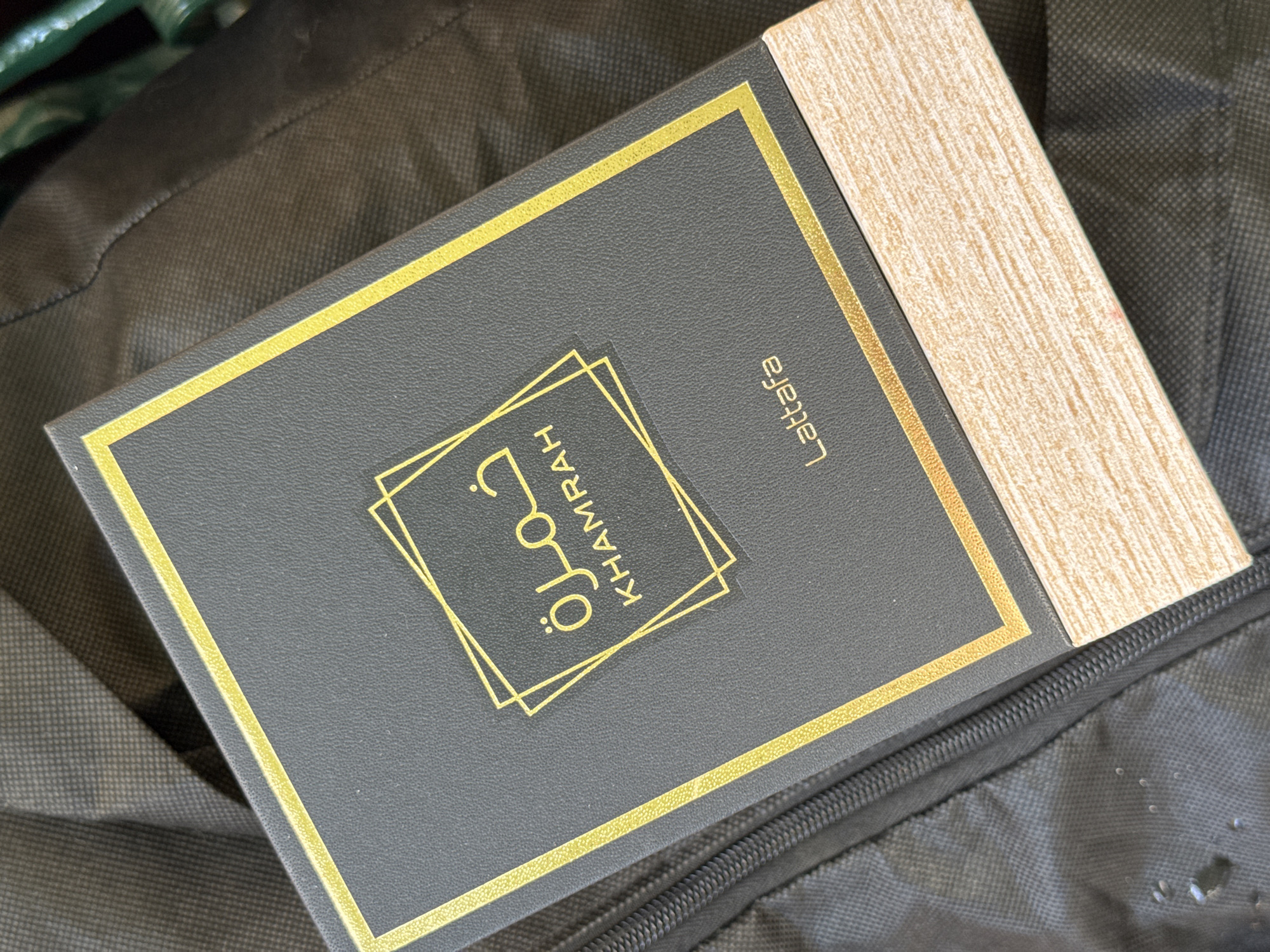 The image shows a close-up, overhead view of a dark gray, rectangular box with gold trim and text. The box is lying on a dark, textured surface that appears to be fabric, possibly a bag or jacket, with a visible zipper on the left side. The top of the box has the Arabic word "خمرة" in gold, above the English word "KHAMRAH" also in gold, enclosed within a geometric gold outline. Below this, the word "Lattafa" is printed in gold on the lower part of the box. The bottom edge of the box has a light wood-grain texture. The scene is devoid of people and there are no contextual clues to suggest a location in Slavyani, Bulgaria.
