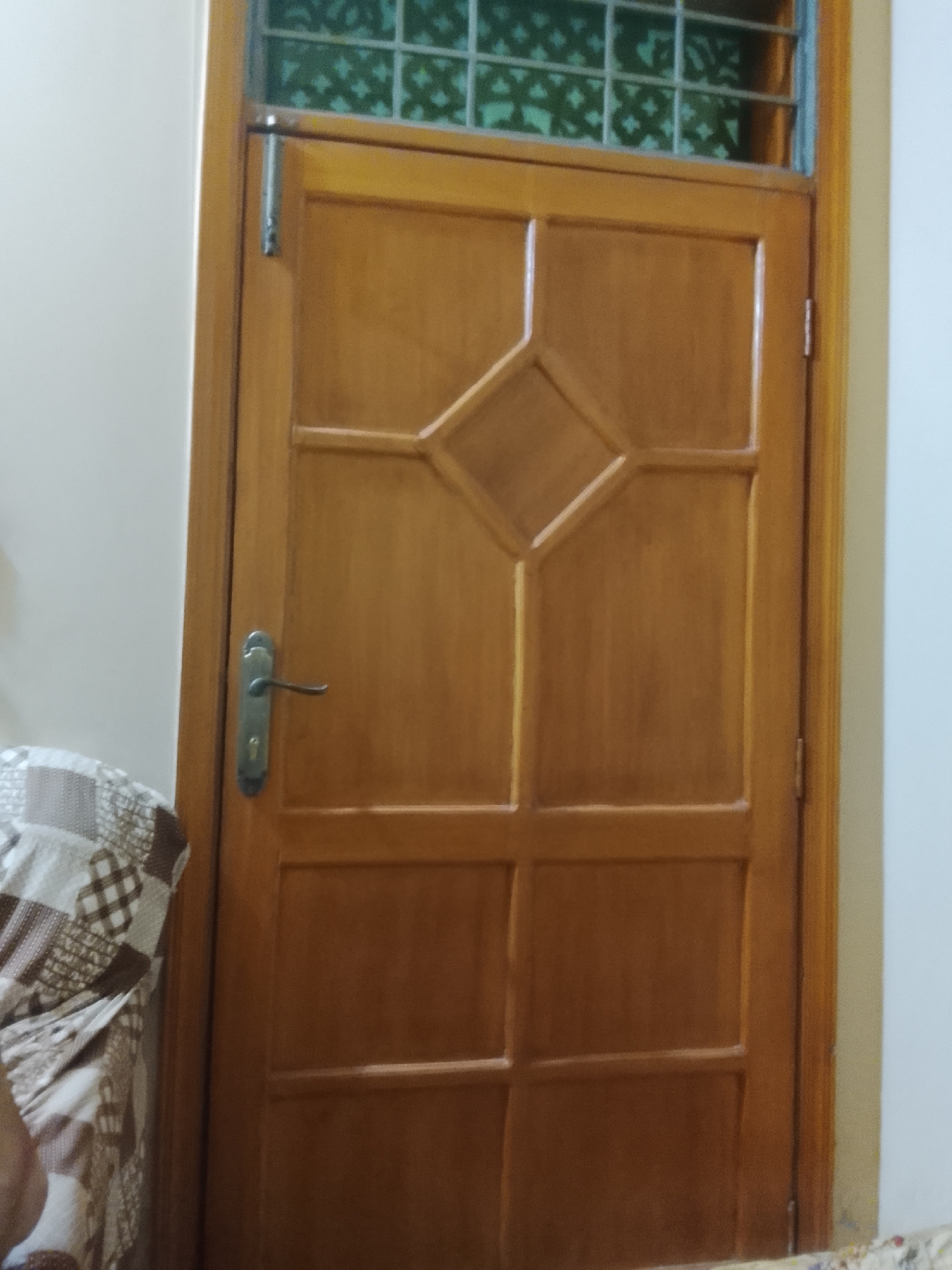 The image shows a wooden door with a diamond-shaped panel near the top. Above the door is a window with a green, patterned glass. A silver door handle is visible on the left side of the door. To the left of the door, a portion of a patterned sofa or chair can be seen. The location is Multan, Pakistan.