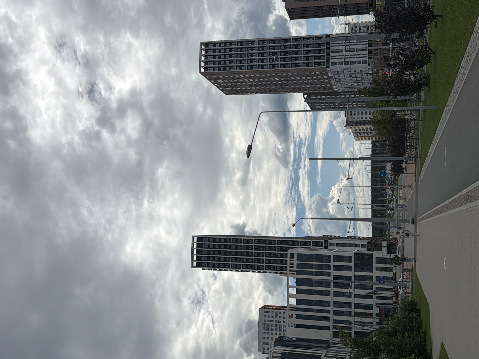 The image shows a street scene with modern, high-rise buildings under a cloudy sky. The buildings are a mix of dark and light facades, with many windows. Streetlights are visible along the street. A paved walkway runs alongside the street, and there is a patch of green grass with trees. Several people are walking on the walkway, dressed casually. The overall impression is that of a contemporary urban environment.
The time of day appears to be daytime, given the brightness of the sky, although the clouds obscure direct sunlight. The weather is overcast, with patches of blue sky and cumulus clouds visible, suggesting a partly cloudy or changeable condition. There are no clear indications of a specific location, though the modern architecture and well-maintained street suggest a developed urban area. The scene is peaceful and uneventful, with people going about their day. No text is clearly visible in the image.