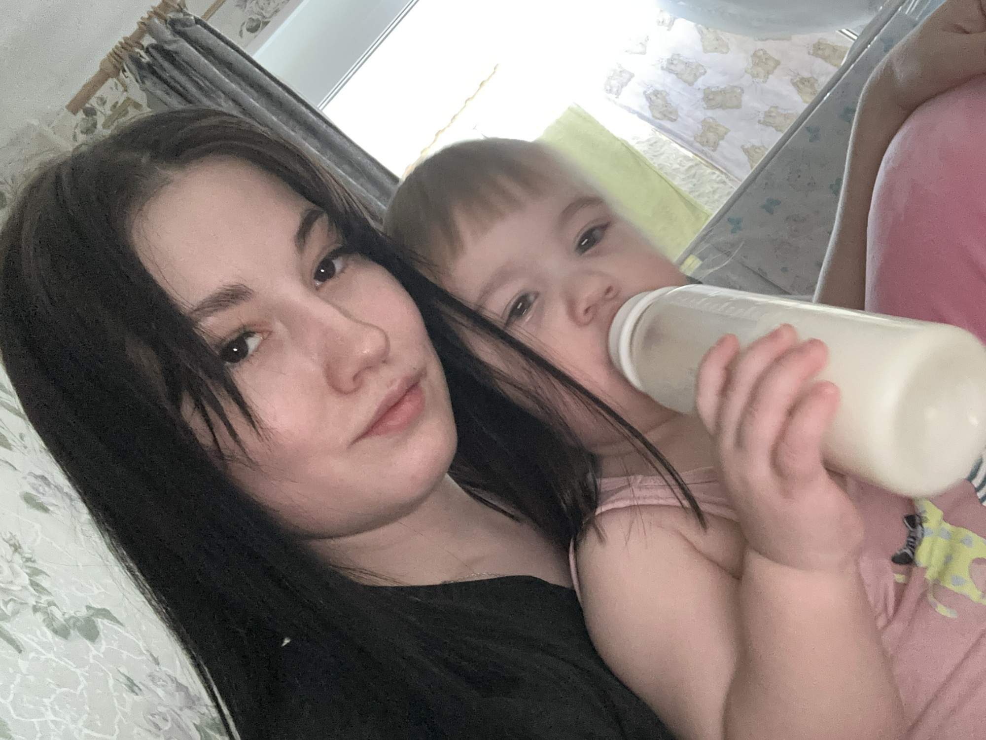 The image is a selfie of a woman with long, dark hair posing with a toddler who is drinking from a baby bottle. The background shows a curtain and bedding with a light-colored floral pattern. The photo appears to be taken indoors in a domestic setting.