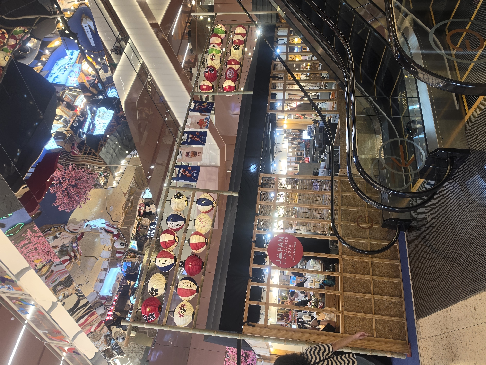 This is an eye-level shot capturing a bustling, multi-story indoor commercial space, likely a shopping mall or food court, featuring a strong Japanese theme. The setting is modern, with reflective surfaces and escalators.
The scene is dominated by vibrant Japanese-style decorations, including numerous red, white, and blue paper lanterns suspended in rows. Many lanterns display Japanese characters such as "ろうしゃい", "こらっしゃい", and various patterns including flowers. Below these, banners resembling traditional "noren" curtains hang, some bearing the text "IZAKAYA" and "JAPAN".
A prominent round red sign reads "JAPAN SIGNATURE 2026" with the Japanese text "ドクトク和食の祭典" (Unique Japanese Food Festival/Celebration) below it, indicating a special event or exhibition. This sign is situated next to a wooden lattice structure, possibly an entrance to a pop-up stall or food vendor area, where people can be seen inside.
An escalator, with sleek black and glass railings, runs diagonally through the right side of the frame, showing movement and the multi-level nature of the building. Mirrored ceiling panels above reflect distorted images of the environment, including artificial cherry blossoms and neon lights, adding to the dynamic and modern aesthetic. People are visible throughout the space, moving around and interacting with the stalls. A person's arm and shoulder are visible in the immediate foreground. The overall ambiance suggests an engaging and festive commercial environment in Bangkok, Thailand.