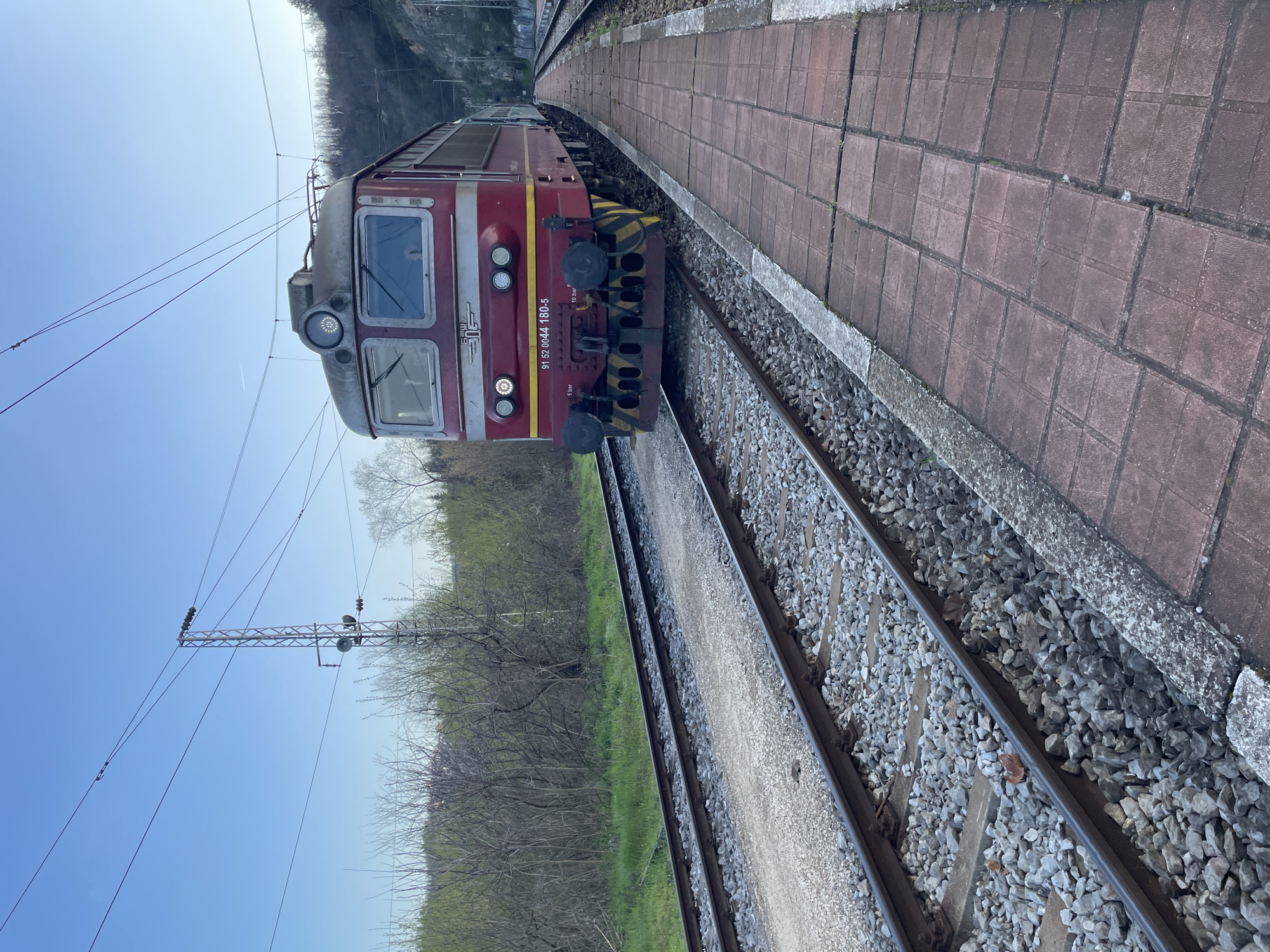 The image shows a red and grey electric locomotive on a curved railway track. The locomotive has a number visible on its front: "91 52 0044 180-5". The track is bordered by grey ballast and a red-brick platform. Across the tracks, green vegetation and trees line the adjacent terrain. In the background, a hillside can be seen. Overhead power lines are present. The location is likely near or within the city of Veliko Tarnovo, Bulgaria, given the number on the locomotive is in a standard format used in Europe.