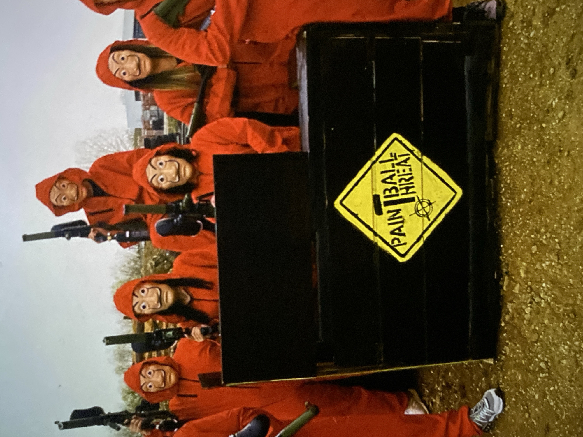 A group of individuals is depicted in an outdoor setting, engaged in a themed paintball activity. At least six participants are visible, all uniformly dressed in bright red hooded jumpsuits. Each person wears a distinctive white Salvador Dalí mask, featuring wide eyes, arched eyebrows, and an iconic upturned moustache, clearly referencing the popular TV series "Money Heist" (La Casa de Papel).
The participants are positioned behind black, box-like barricades or bunkers, holding paintball markers (guns) in their hands, suggesting strategic play within the game. They appear to be in a formation, with some individuals taking cover in the foreground and others extending into the background.
A prominent yellow diamond-shaped sign is affixed to one of the black barricades in the foreground. The sign bears the black text "PAIN BALL THREAT," with "PAIN" on the first line and "BALL THREAT" on the second, and a target symbol beneath, explicitly indicating the nature of the activity.
The scene unfolds during the daytime, under what appears to be an overcast sky, providing soft, diffused lighting without strong shadows. The ground is a rough, yellowish-brown surface, likely dirt or gravel, typical of an outdoor recreational area. Faint, indistinct outlines of distant trees or structures are visible against the light sky in the background. A foot clad in a grey and white sneaker is partially visible in the bottom right corner. The overall image captures a moment of active, themed paintball play, set in an open outdoor environment. No specific visual cues within the image identify the location as Bankya, Bulgaria, although this is provided as context.
