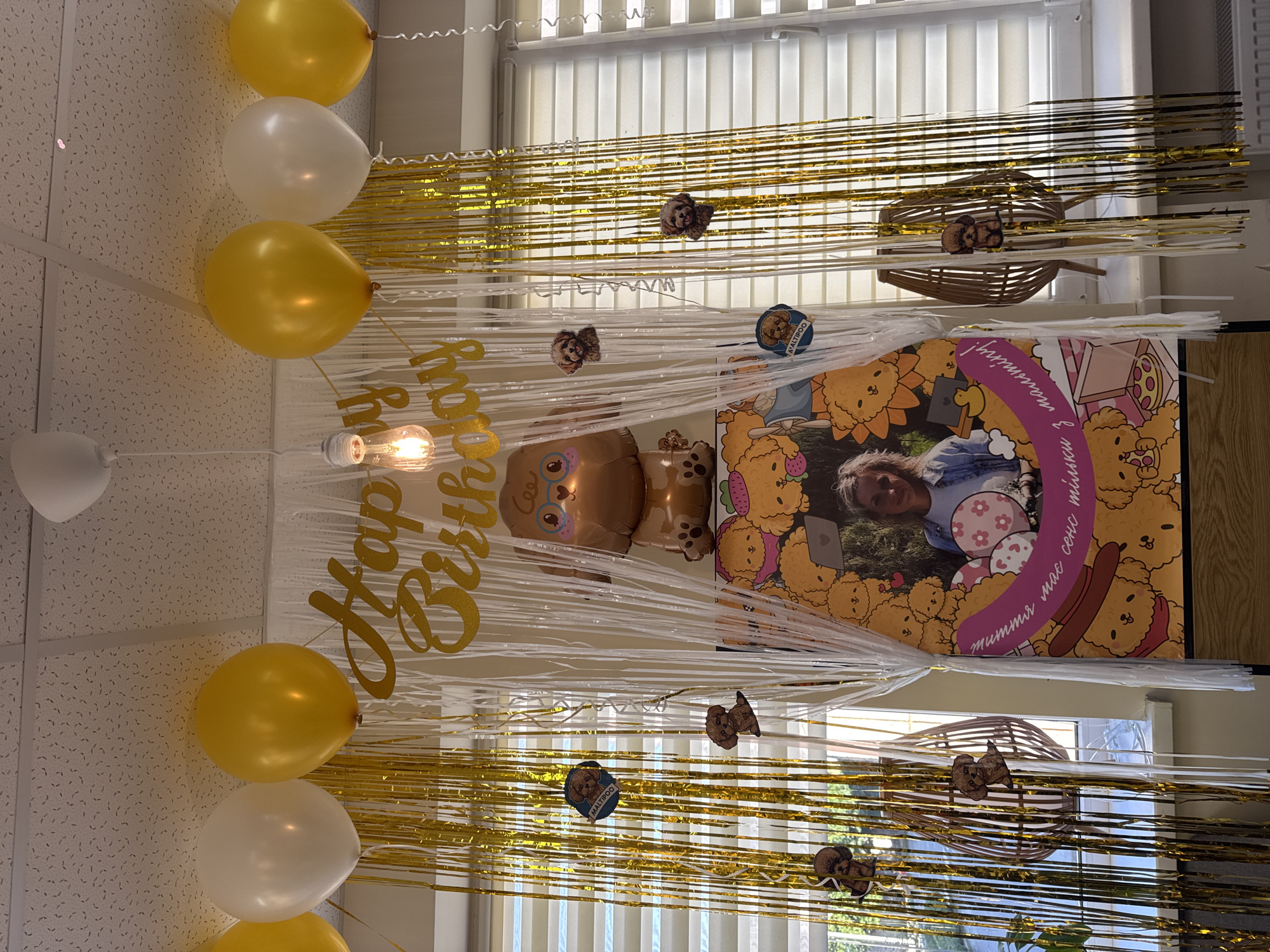 This image depicts a birthday decoration setup indoors, likely in a room with windows. The scene is adorned with gold and white streamers and balloons in yellow and white. Prominently displayed is a large banner featuring cartoon dogs and a photograph of a woman. There is also a large dog-shaped balloon. Several smaller dog cutouts are attached to the streamers. A hanging lightbulb is visible at the top center of the frame. The overall ambiance suggests a festive, celebratory atmosphere. The location is identified as Kremenchuk, Ukraine.