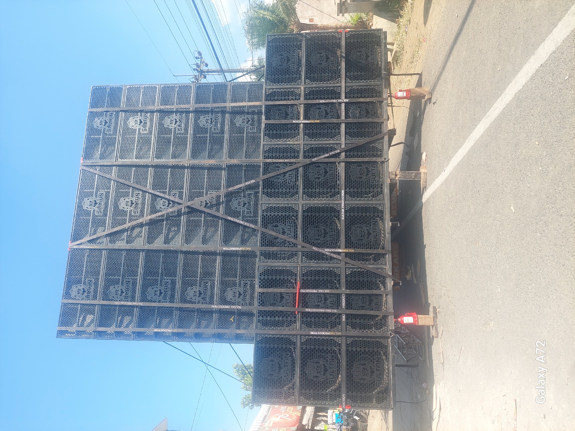 The image shows a large stack of black speaker cabinets labeled "SULTAN PRODUCTION" on a flatbed truck. The cabinets are arranged in multiple tiers and are secured with black straps. The truck is parked on a paved road, likely in Sengguruh, Indonesia, given the context. The background shows a clear blue sky, some power lines, a building on the left with signage, and vegetation on the right. A red jack is visible supporting the rear of the truck. There are no people clearly visible in the frame. The overall scene suggests preparation for an event or performance, with the sound system being transported or set up.