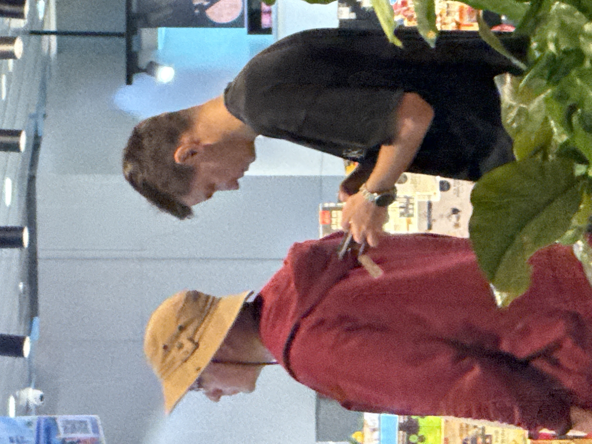 Two individuals are visible in a brightly lit indoor environment, likely a retail setting, with shelves and displays in the background. On the left, a person wearing a tan bucket hat and a dark red hooded jacket is facing towards the right, with their hands near their chest. On the right, a person with short dark hair, wearing a black t-shirt, is looking down at their hands which are holding a mobile device. Their left wrist is adorned with a wristwatch. The scene appears to be in Bắc Cam Ranh, Vietnam, given the context.