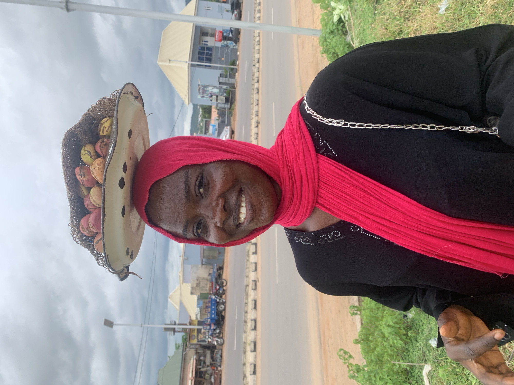 A medium shot captures a Black woman with dark skin, wearing a bright pink hijab and a black abaya with beaded detailing around the neckline, in Adankolo, Nigeria. She is smiling broadly, revealing her teeth, and has a tray of various fruits, including what appear to be coconuts, balanced on her head. The tray is white with a dark rim and a woven material lining. She is looking directly at the camera. Her left hand is visible, extended slightly towards the viewer. Behind her, a paved road runs horizontally, with a sidewalk or median strip separating it from the grassy shoulder where she stands. In the background, buildings with light-colored roofs and facades, streetlights, and parked vehicles, including motorcycles and a blue truck, are visible. The sky is overcast with gray clouds.