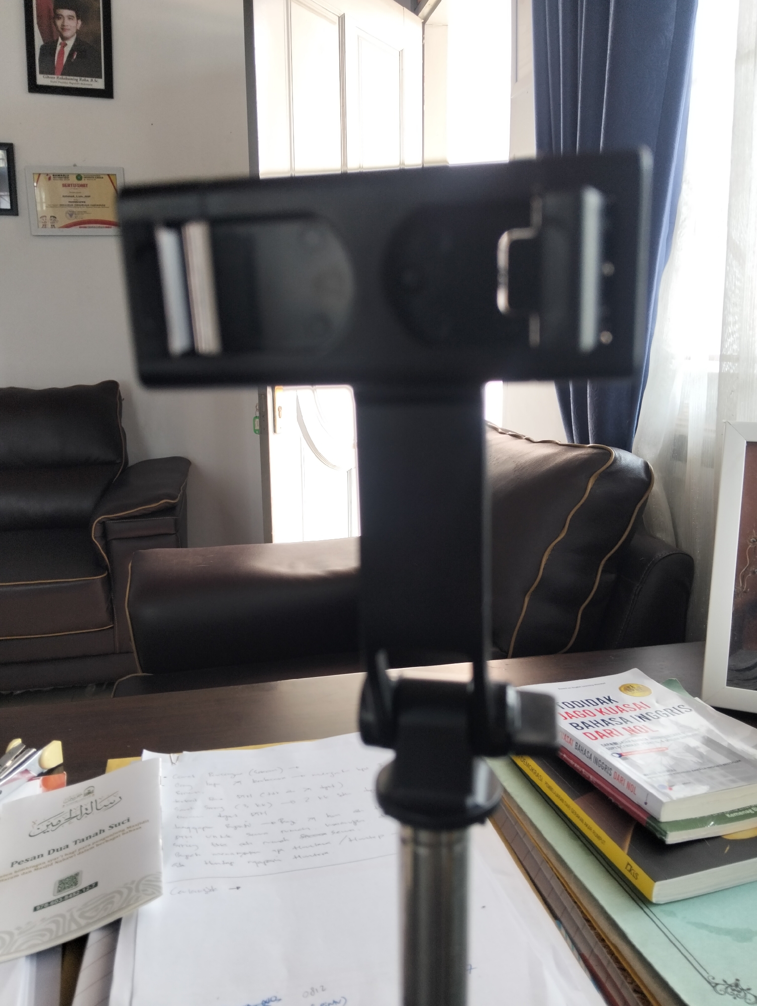 A cellphone tripod is in the foreground, with a portion of a dark leather sofa and a dark wooden desk visible behind it. On the desk are several books, one titled "GOIDAK JAGO KUASAI BAHASA INGGRIS DARI NOL," and a white booklet with Arabic script. A framed photograph of a man in a suit and tie hangs on the wall to the left, alongside a framed certificate. A doorway with a light-colored door is visible in the background, partially obscured by a blue curtain and white sheer curtains. The setting appears to be an office or living room.